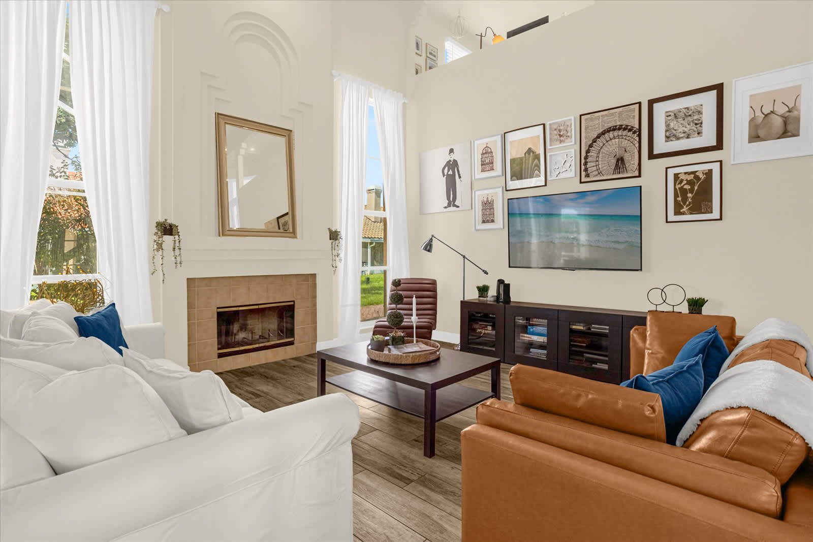 A bright living room features a white slipcovered sofa and a brown leather sofa, both with blue throw pillows, facing a dark wood coffee table. A large flat-screen TV is mounted above a media console, and a fireplace with a decorative mirror above it adds a focal point.