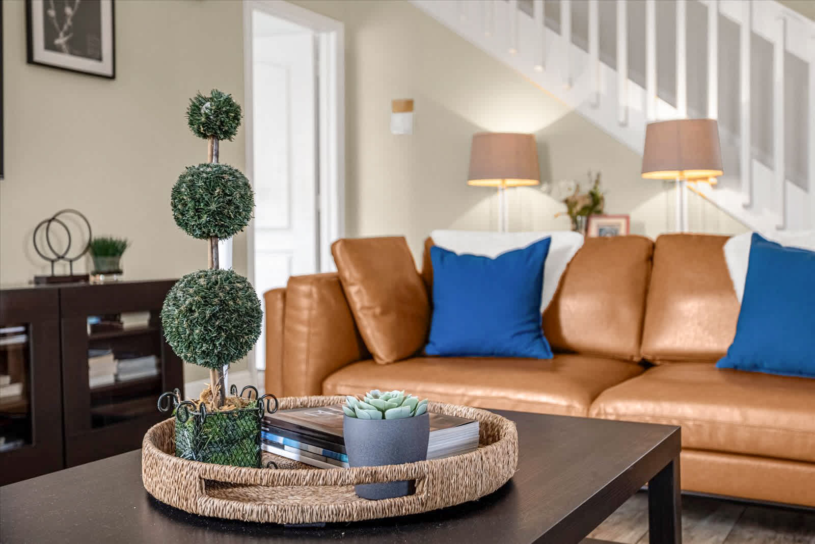 Cozy living room featuring a tan leather sofa with blue accent pillows, a dark wood coffee table holding a woven tray with decor, and a tall topiary plant, with a staircase visible in the background.