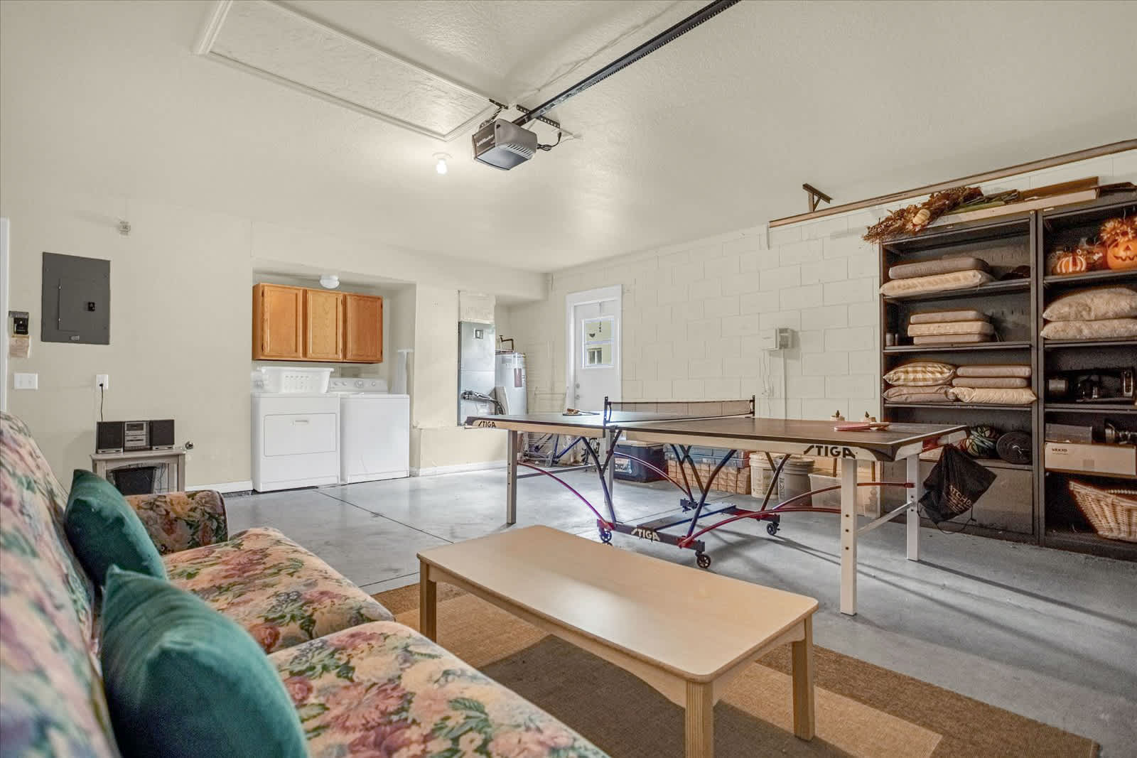 Spacious game room with a concrete floor, featuring a ping-pong table in the center and a floral-patterned sofa on the left. A laundry area with a washer, dryer, and wooden cabinets is visible in the background, alongside a shelving unit filled with various items.