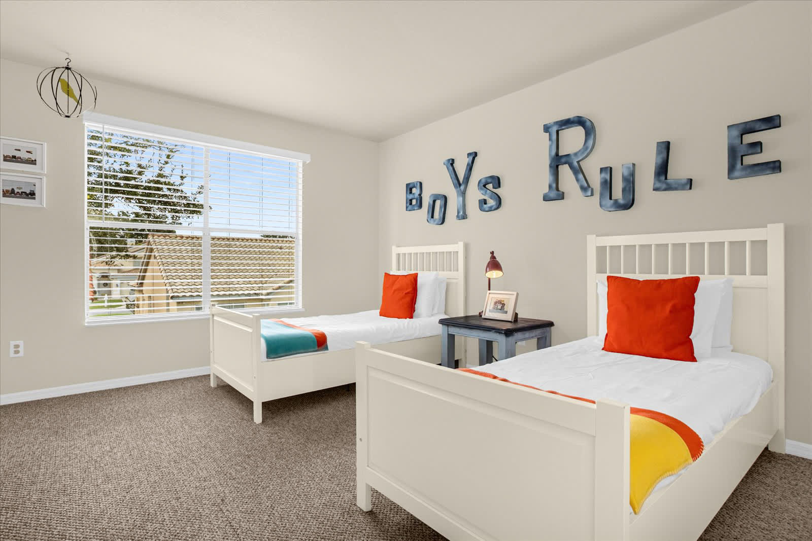 Bright boys' bedroom featuring two white twin beds with orange pillows and colorful throws, set against a light beige wall adorned with large 'BOYS RULE' lettering. A window with blinds overlooks the exterior, and a small nightstand with a lamp sits between the beds on a carpeted floor.