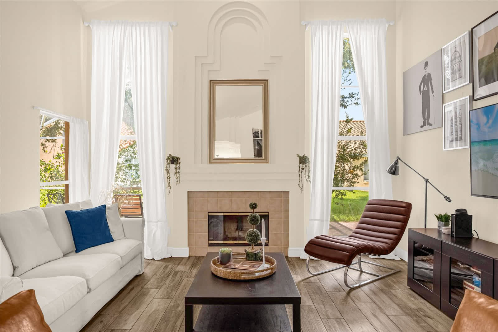 Spacious living room with high vaulted ceilings, a white sofa, a brown leather lounge chair, a dark coffee table, and a fireplace with a mirror above, flanked by large windows with white sheer curtains.