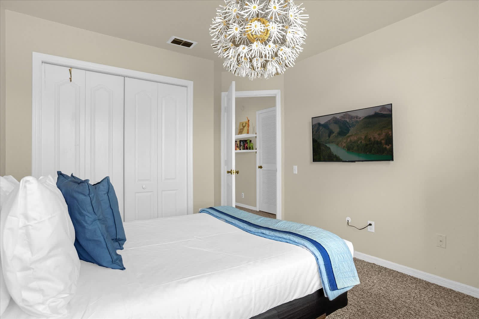 Another angle of the bedroom showing a wall-mounted TV, white bi-fold closet doors, and a glimpse into an adjacent room through an open door. The dandelion-like chandelier is prominent, and the room has beige walls and carpet.
