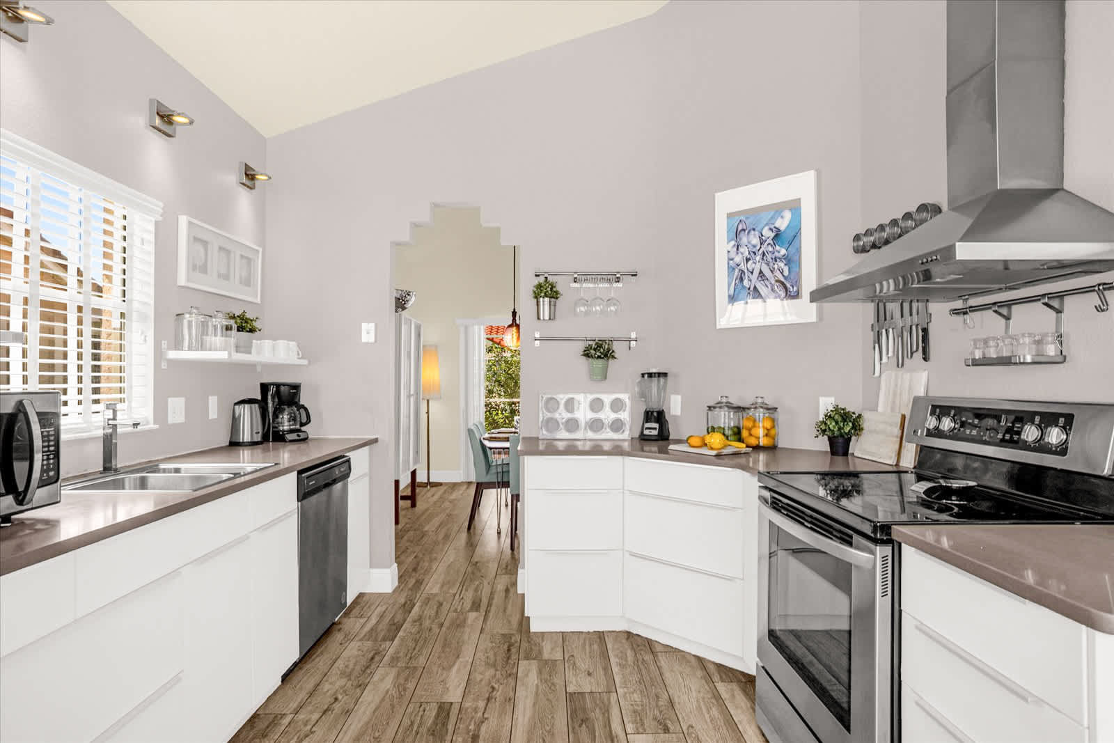 Modern kitchen with white flat-panel cabinets, dark gray countertops, and stainless steel appliances including a range, range hood, and dishwasher, all set on wood-look tile flooring.