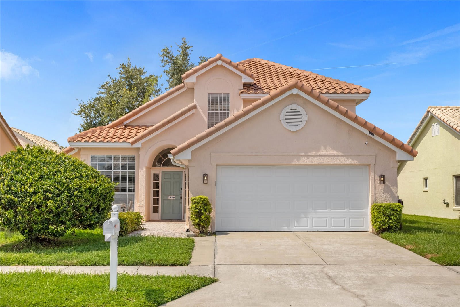 The front exterior of a two-story house features a light peach stucco facade with a red tile roof, a white garage door, and a light blue front door. Lush green bushes flank the entrance, and a mailbox stands by the driveway under a clear blue sky.