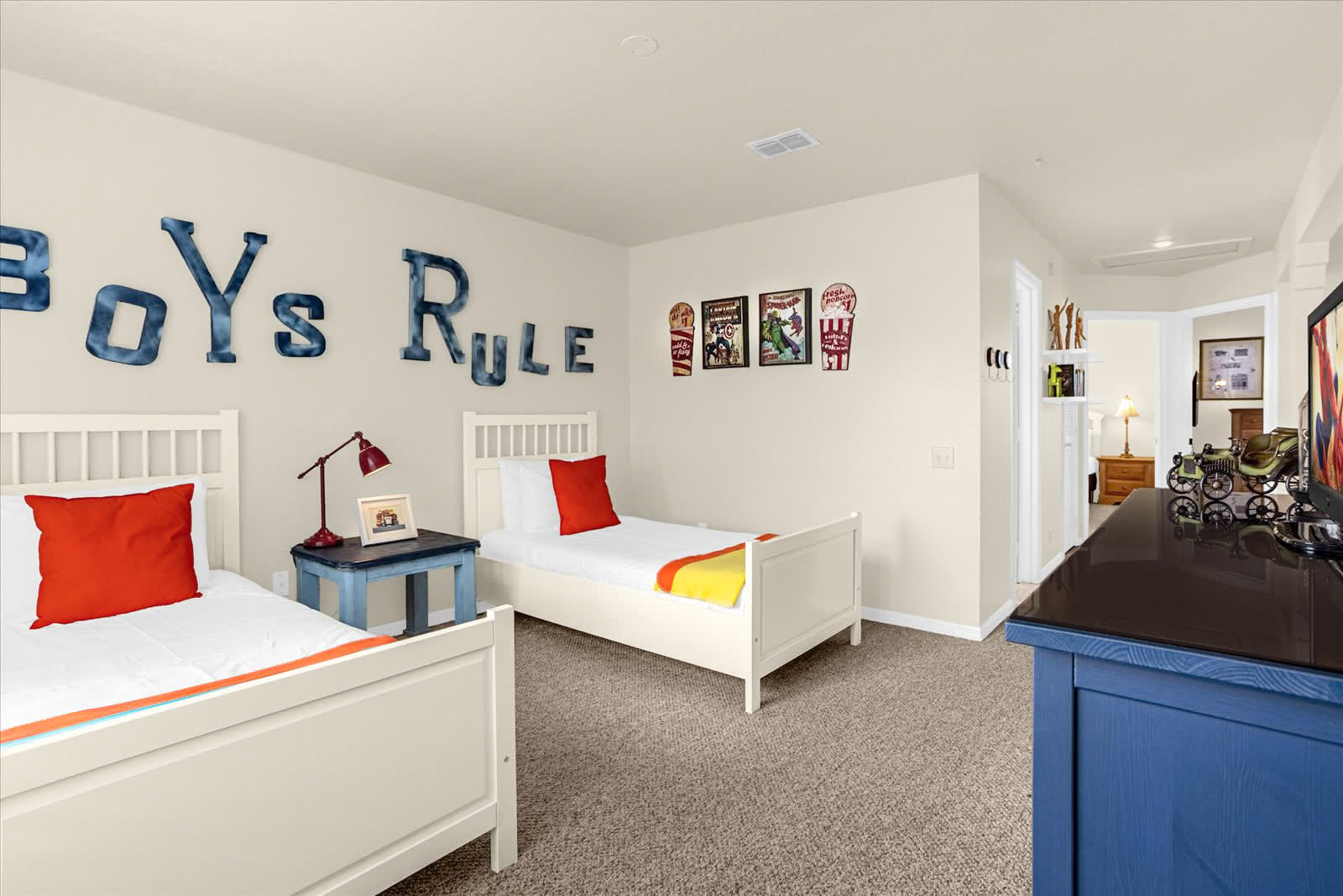 Another view of the boys' bedroom, showcasing the two white twin beds with orange pillows and a blue nightstand with a lamp. The 'BOYS RULE' wall decor is prominent, and a dark blue dresser with a TV and framed comic book art above it is visible on the right.
