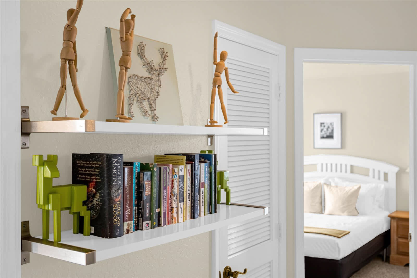 A white wall-mounted bookshelf filled with books and decorative wooden figures, positioned in a hallway or transition space. A bright bedroom with a white headboard is visible through an open doorway in the background.