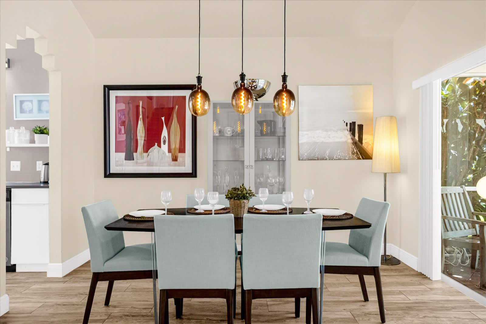 View of the dining room with a dark wood table and light blue chairs, three pendant lights, and artwork, showing an arched doorway leading to the kitchen and a sliding glass door to an outdoor area.