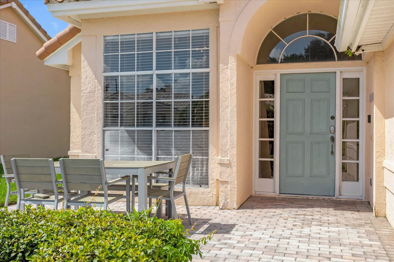 Inviting front entrance of a stucco house with a light blue door featuring an arched transom window and sidelights. A large window with blinds is to the left, and an outdoor dining set with a grey table and chairs sits on a paved patio area, surrounded by green landscaping.