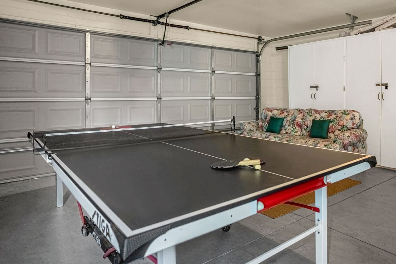 A garage converted into a game room features a black Stiga ping pong table with paddles and balls, set against a gray paneled garage door. A floral patterned sofa provides seating, and white storage cabinets line one wall.