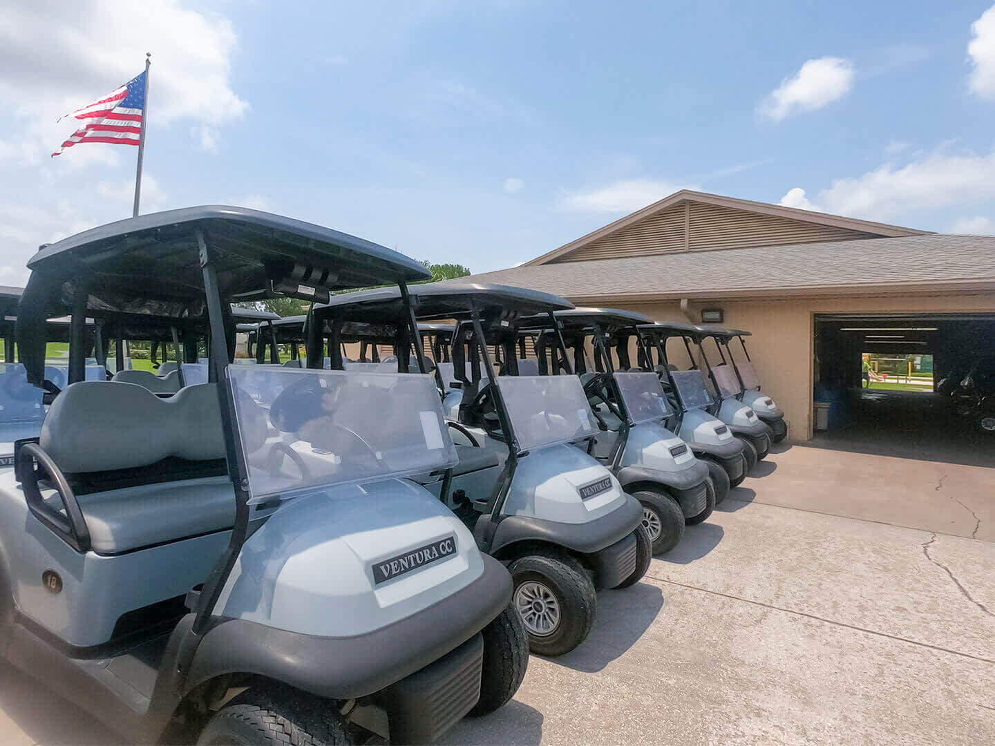 A line of light grey golf carts, branded 'VENTURA CC', parked neatly outside a clubhouse building under a clear blue sky with an American flag waving. The scene suggests a golf course or resort setting, ready for guests.