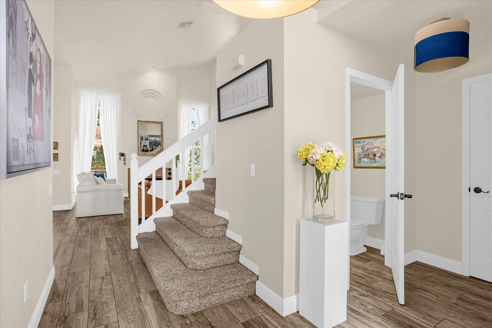 Bright foyer with wood-look tile flooring, a carpeted staircase with white railings, a decorative pedestal with a vase of flowers, and an open doorway revealing a partial bathroom.