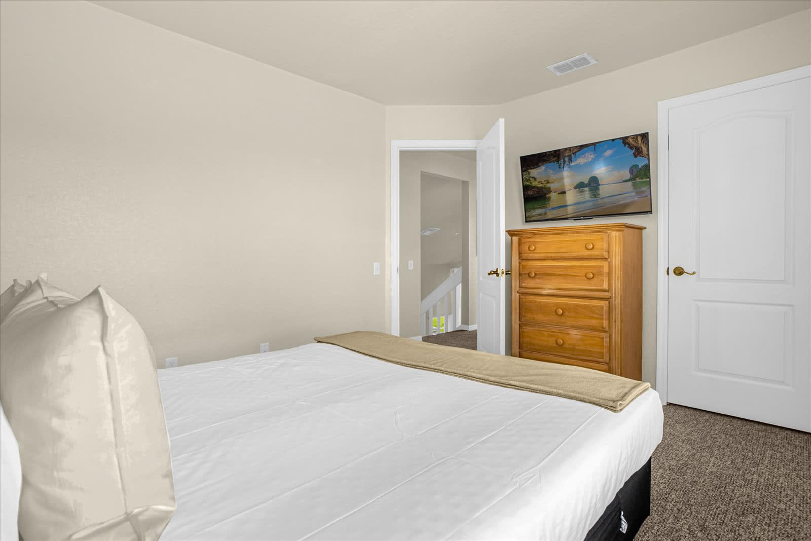 Another perspective of the bedroom, showcasing a wall-mounted TV above a wooden dresser, and an open doorway leading to a staircase. The queen bed with a white headboard is visible, and the room has beige walls and carpet.