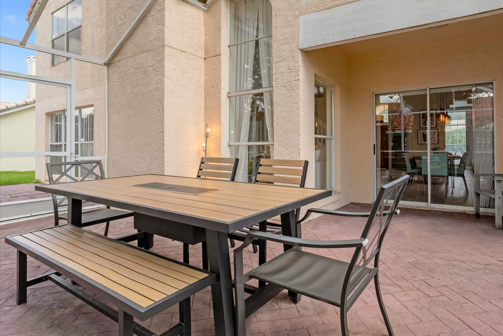A wide shot of a screened patio shows a modern outdoor dining set with a table, bench, and chairs on red brick pavers, with a gas grill nearby. Large glass sliding doors connect the patio to the interior dining area, and the space is surrounded by lush greenery.