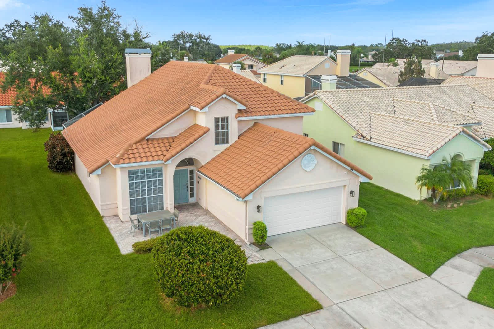 Aerial view of a single-family home with a terracotta tile roof and a light-colored stucco exterior, featuring an attached two-car garage and a paved driveway. The property includes a well-maintained green lawn and an outdoor dining area at the front, surrounded by similar residential homes.