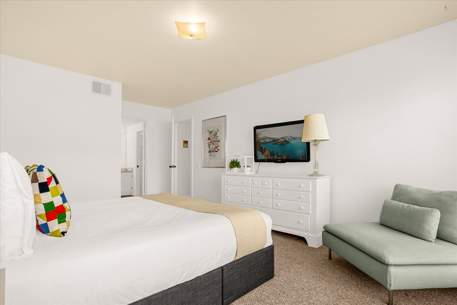 Another view of the bright bedroom, showing a wall-mounted TV above a white dresser, a light green accent chair, and a glimpse of a doorway leading to a bathroom. The room features white walls and beige carpet.