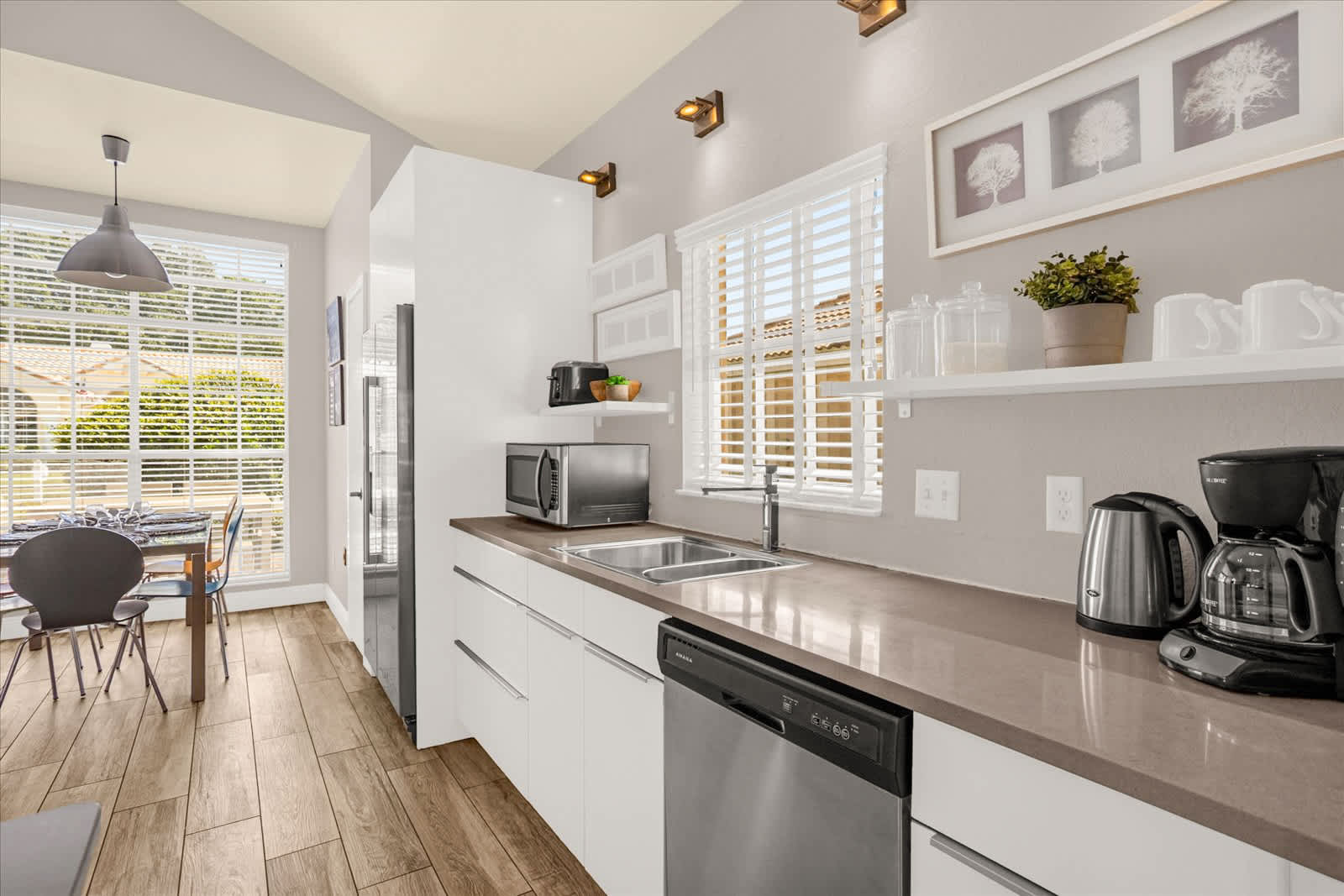 Kitchen area with white cabinets, dark countertops, a stainless steel sink, microwave, and coffee maker, with a window featuring white blinds and open shelving, leading to a breakfast nook.