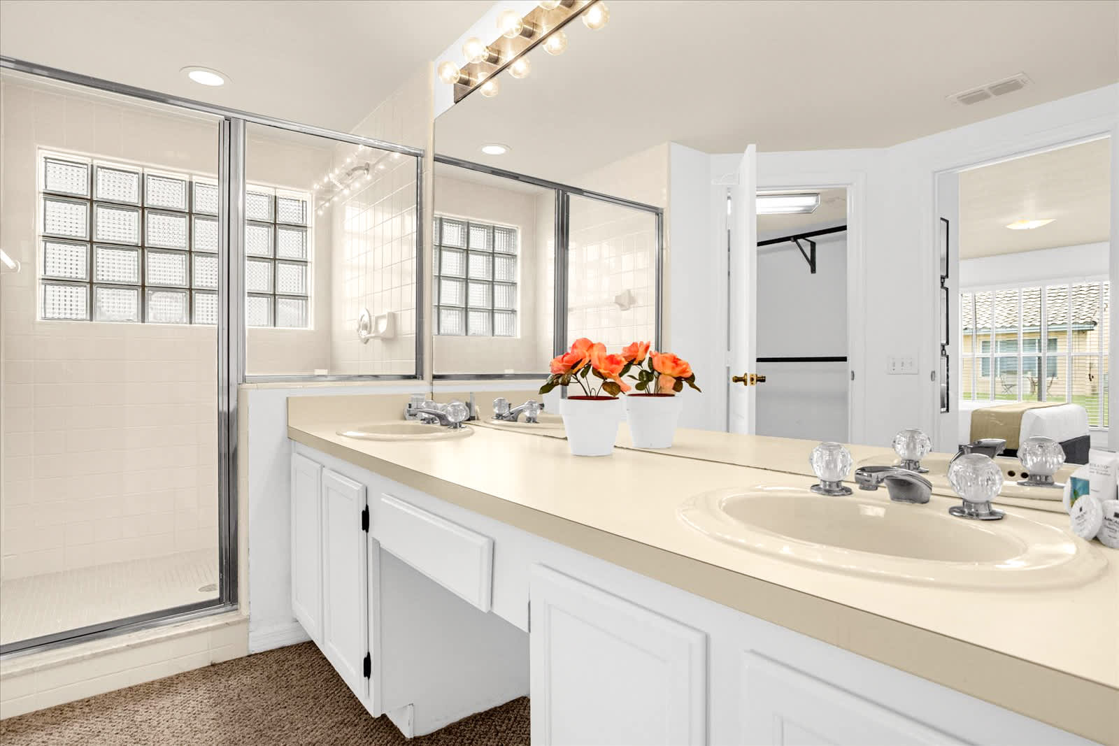 Close-up view of the double vanity with light countertops and white cabinets, featuring two sinks and a large mirror, adjacent to a glass-enclosed shower with glass block windows. The floor is carpeted.