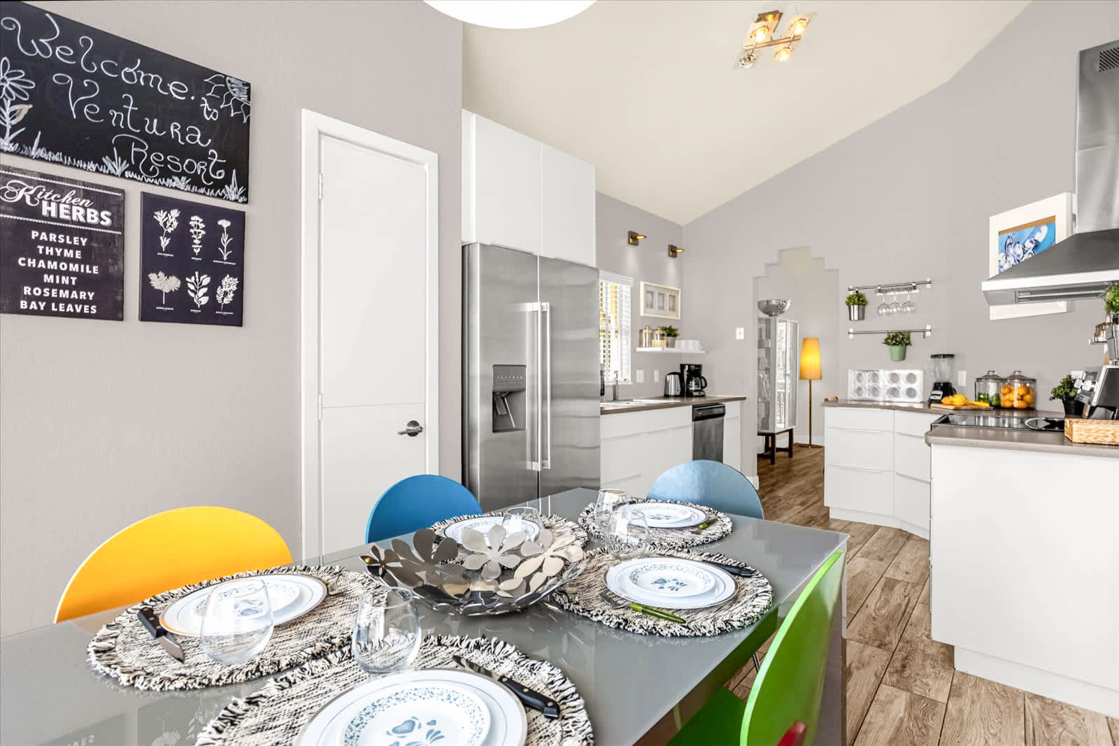 Breakfast nook area with a gray table and colorful chairs, adjacent to the kitchen with a large stainless steel refrigerator and a decorative chalkboard sign on the wall.