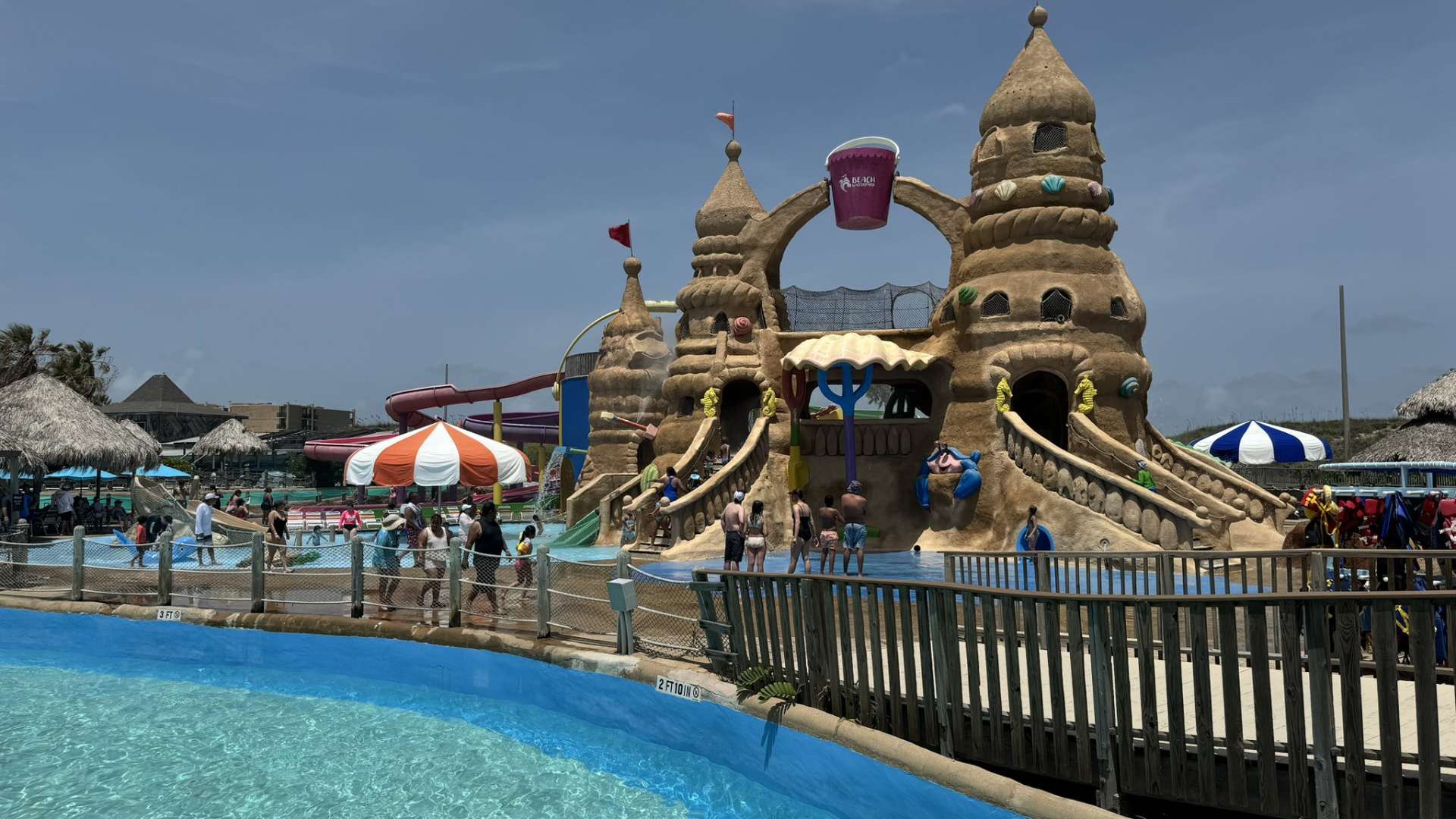 A vibrant water park scene featuring a large sandcastle-themed play structure with slides, a tipping bucket, and multiple pools, bustling with people enjoying the sunny day. A blue lazy river or shallow pool is in the foreground, separated by a wooden fence.