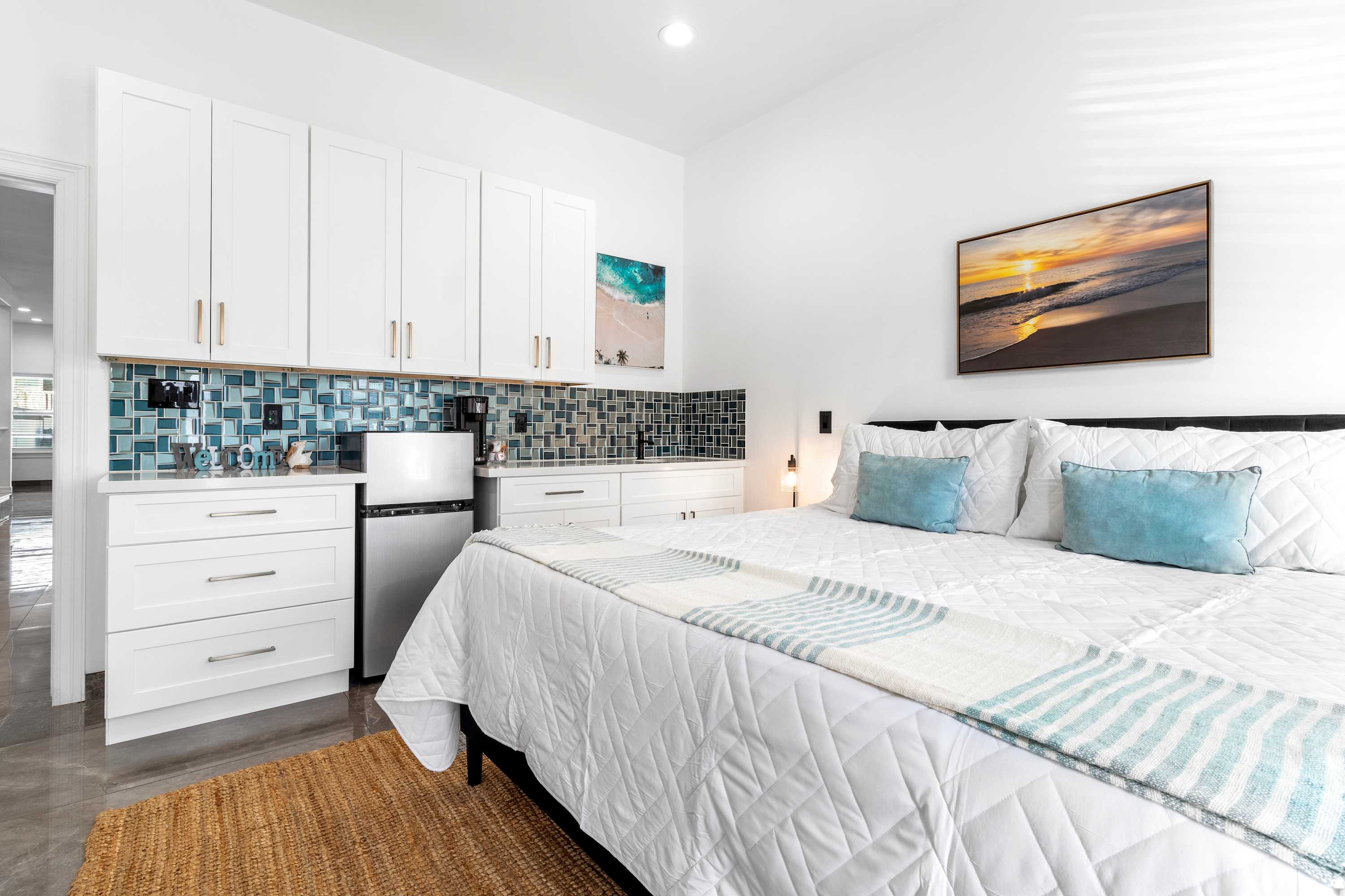 Bright bedroom featuring a white quilted king-size bed with blue accent pillows, a built-in white kitchenette with blue mosaic tile backsplash, and a large ocean-themed artwork above the bed.
