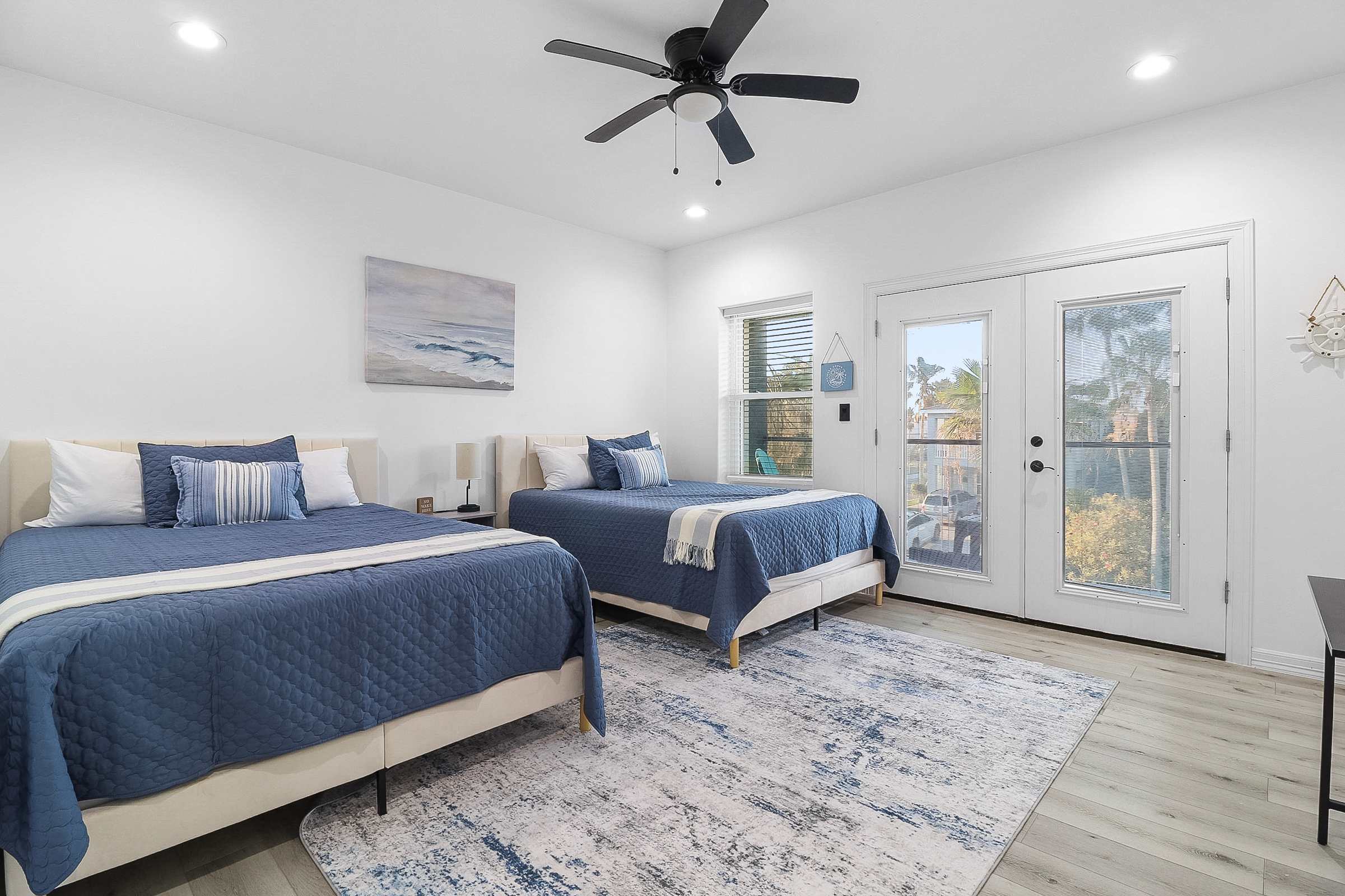 A spacious bedroom with two queen beds dressed in blue quilted bedding, a large blue and white abstract rug, light wood-look flooring, and a glass-paned door leading to an outdoor area.