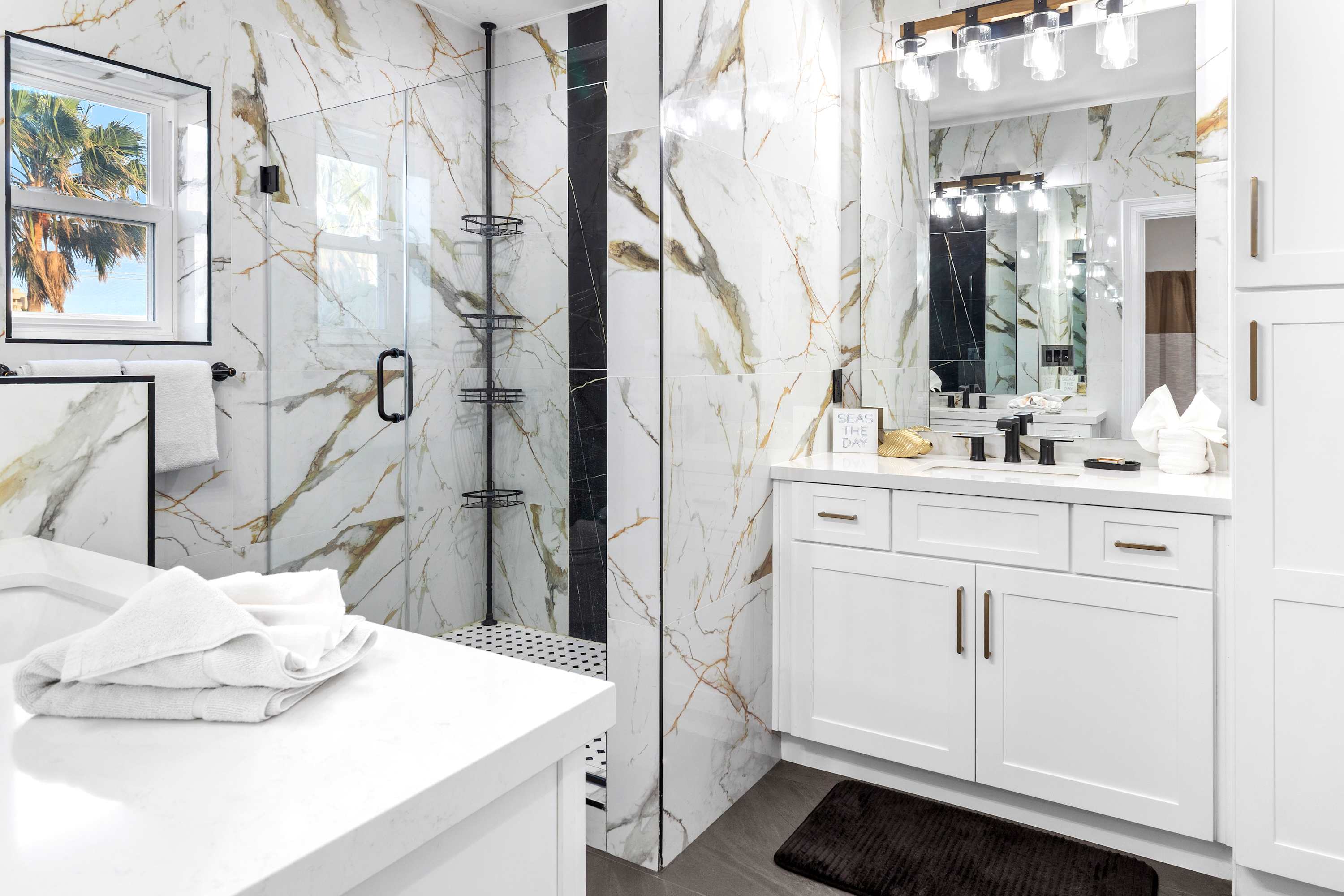 Full view of the elegant bathroom, showcasing the white vanity, large mirror, glass shower enclosure, and a window providing natural light, all surrounded by dramatic marble-look tile.