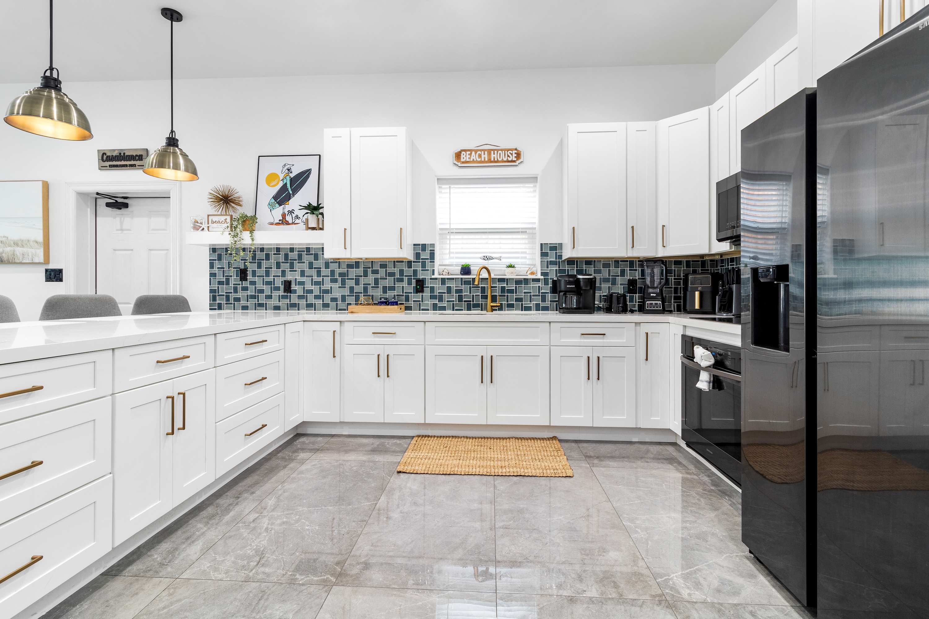 Well-lit kitchen featuring white shaker cabinets, dark grey appliances, and a striking blue mosaic tile backsplash. A "Beach House" sign hangs above the window, adding to the coastal theme.