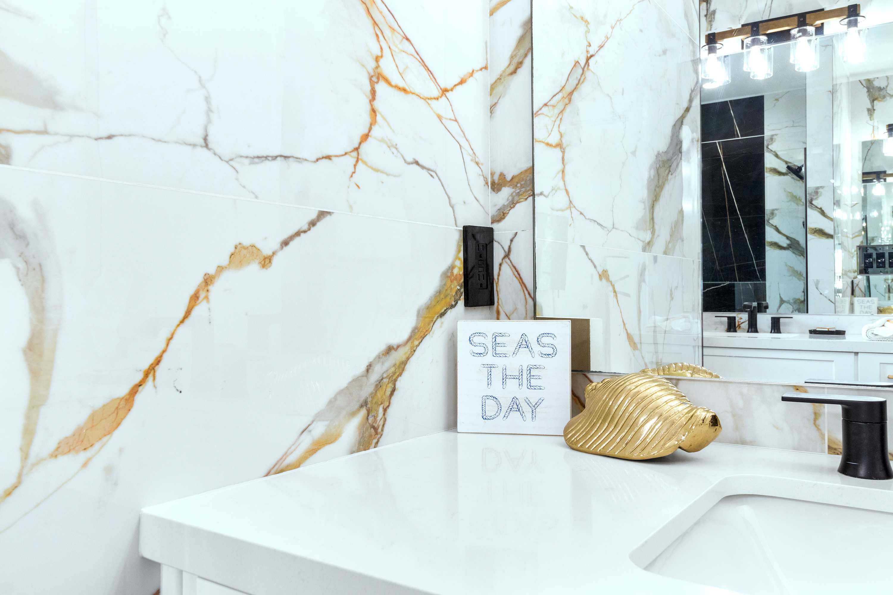 Close-up of the bathroom vanity, featuring a white quartz countertop, black faucet, and a decorative "SEAS THE DAY" sign with a gold shell, set against the striking marble-look tile.