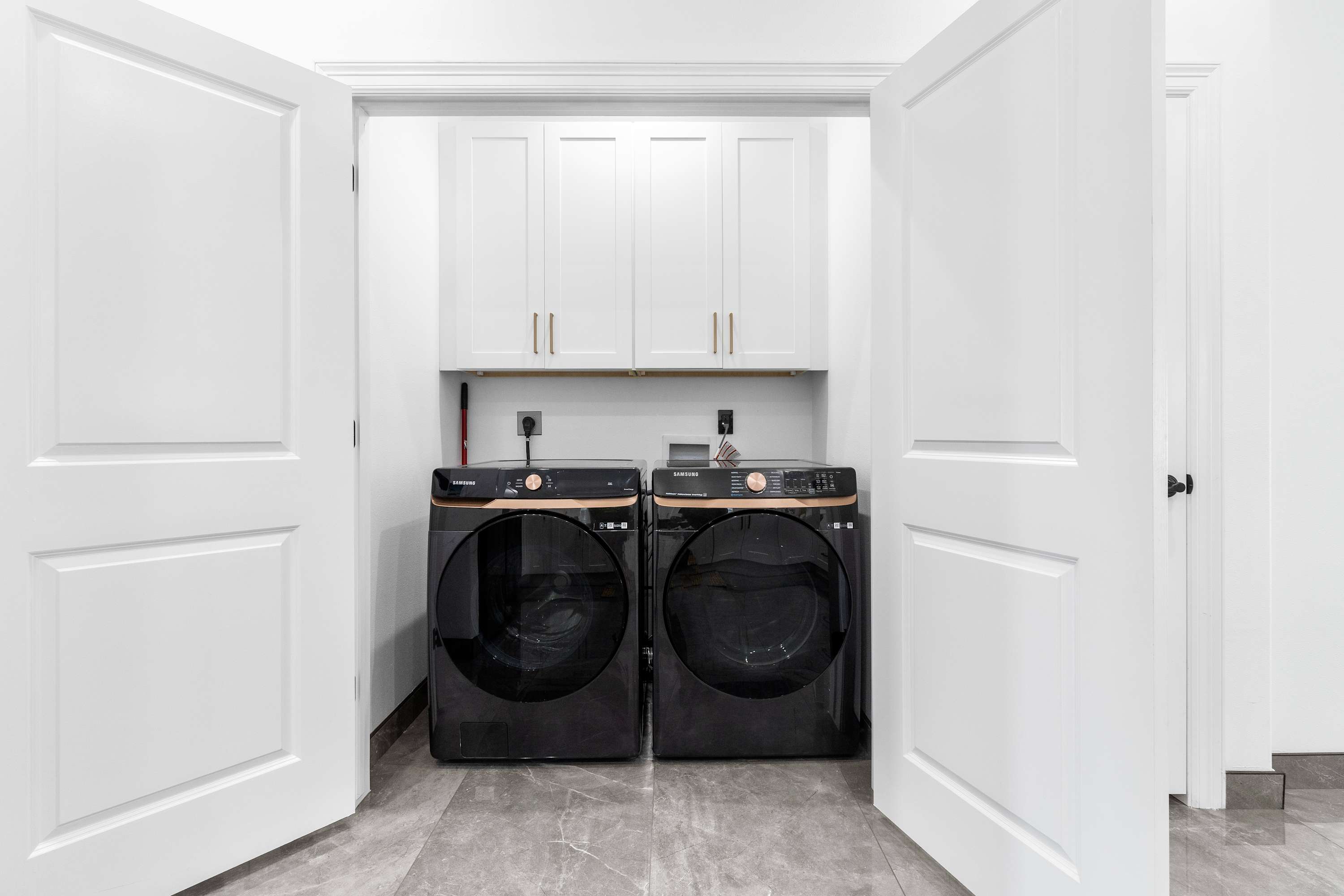 A dedicated laundry closet with a black Samsung washer and dryer set, white upper cabinets with gold handles, and dark grey tiled flooring, enclosed by white bi-fold doors.