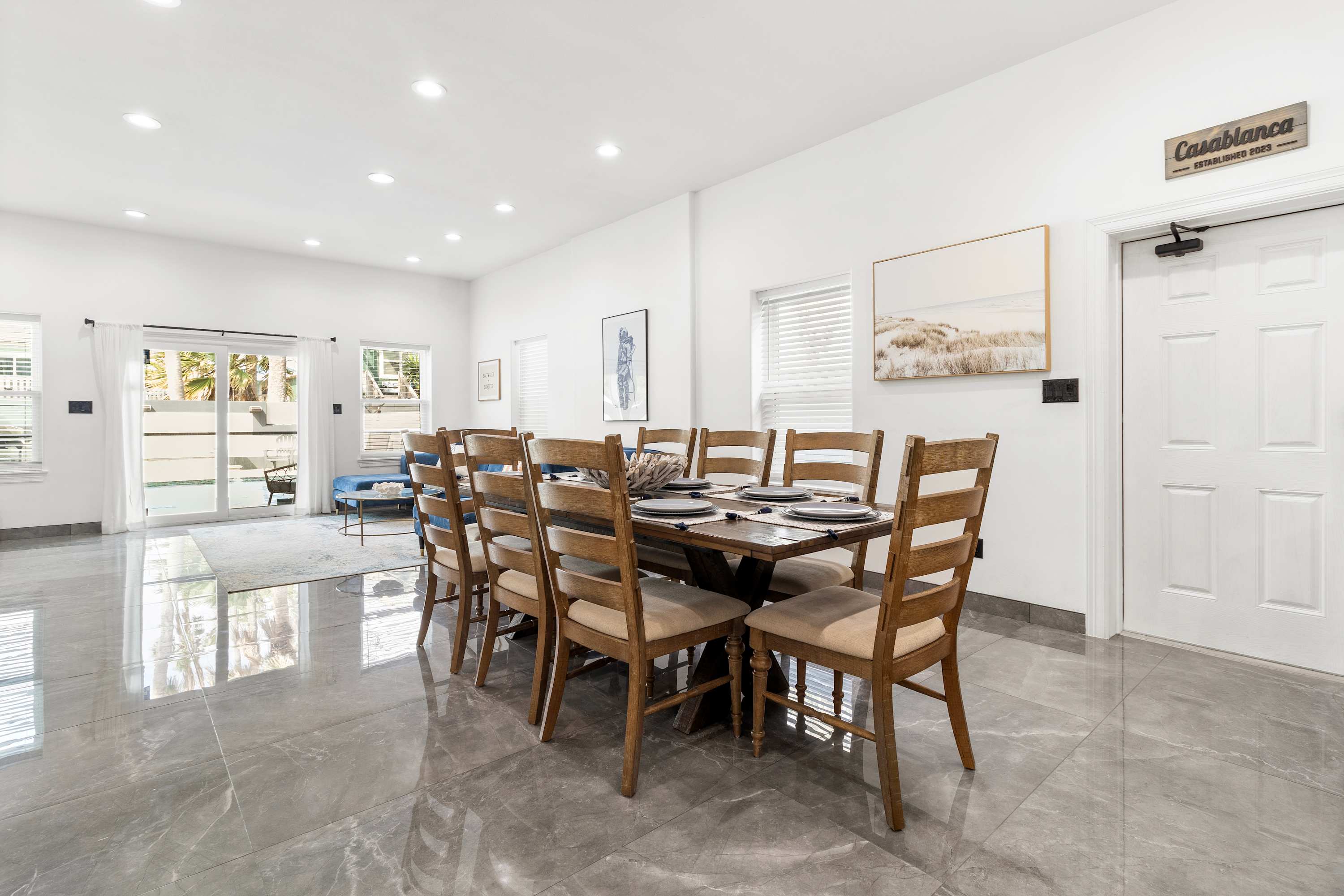 Bright dining area with a large wooden table and eight chairs, illuminated by natural light from multiple windows. Sliding glass doors lead to an outdoor patio, and a white door with a "Casablanca" sign is visible.