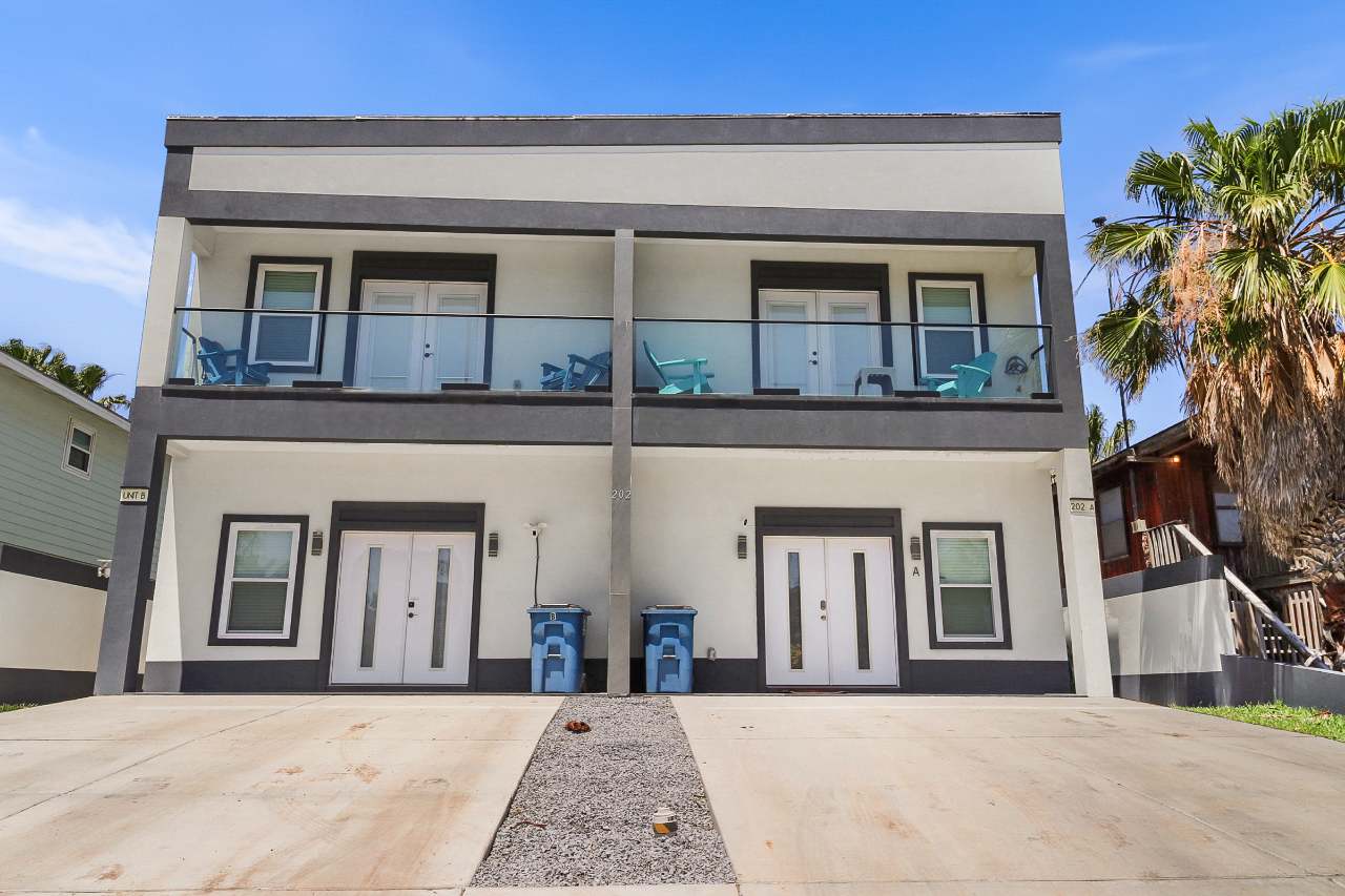 The front exterior of a modern two-story townhouse building with a white and dark grey facade, featuring multiple balconies with glass railings, white entry doors, and a concrete driveway.
