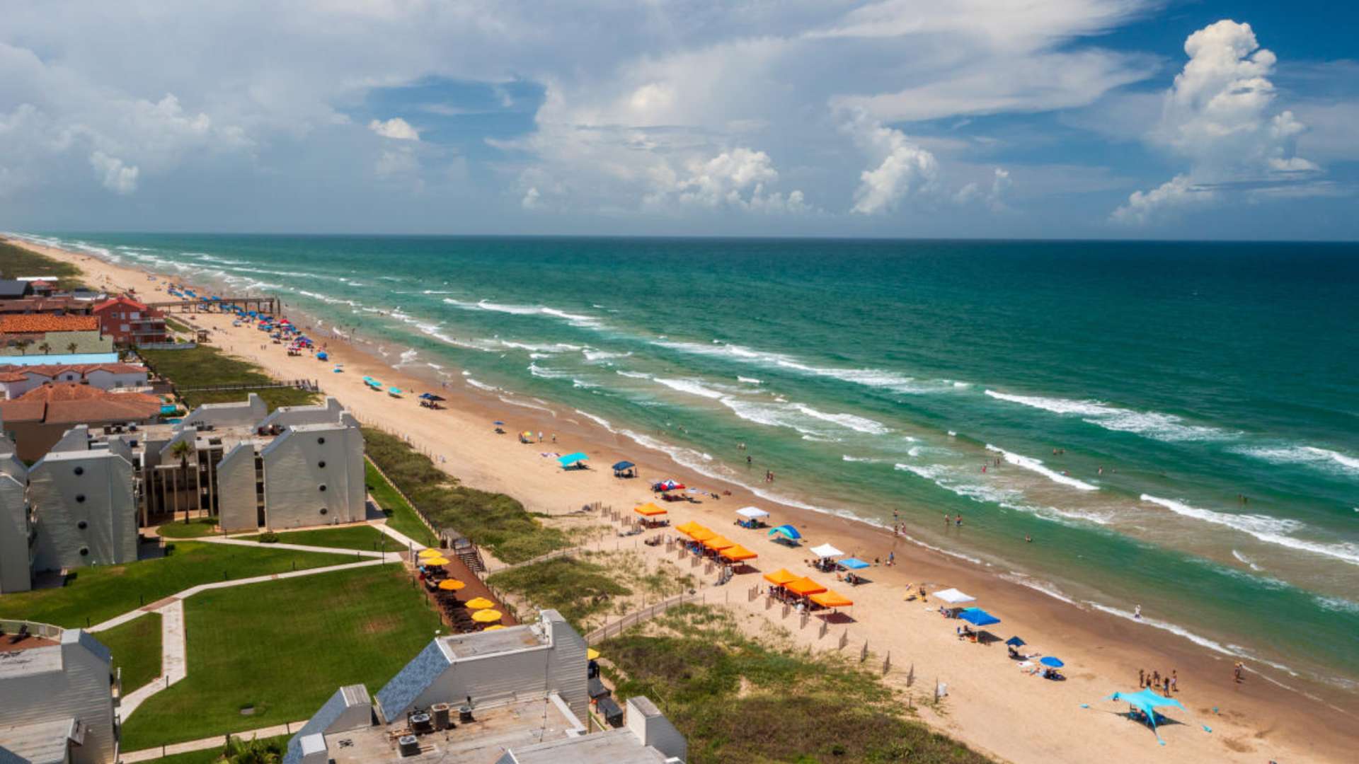 An expansive aerial view of a sandy beach stretching along a vibrant turquoise ocean with gentle waves, dotted with numerous colorful beach umbrellas and chairs. Modern white buildings with green lawns line the coast under a partly cloudy sky.