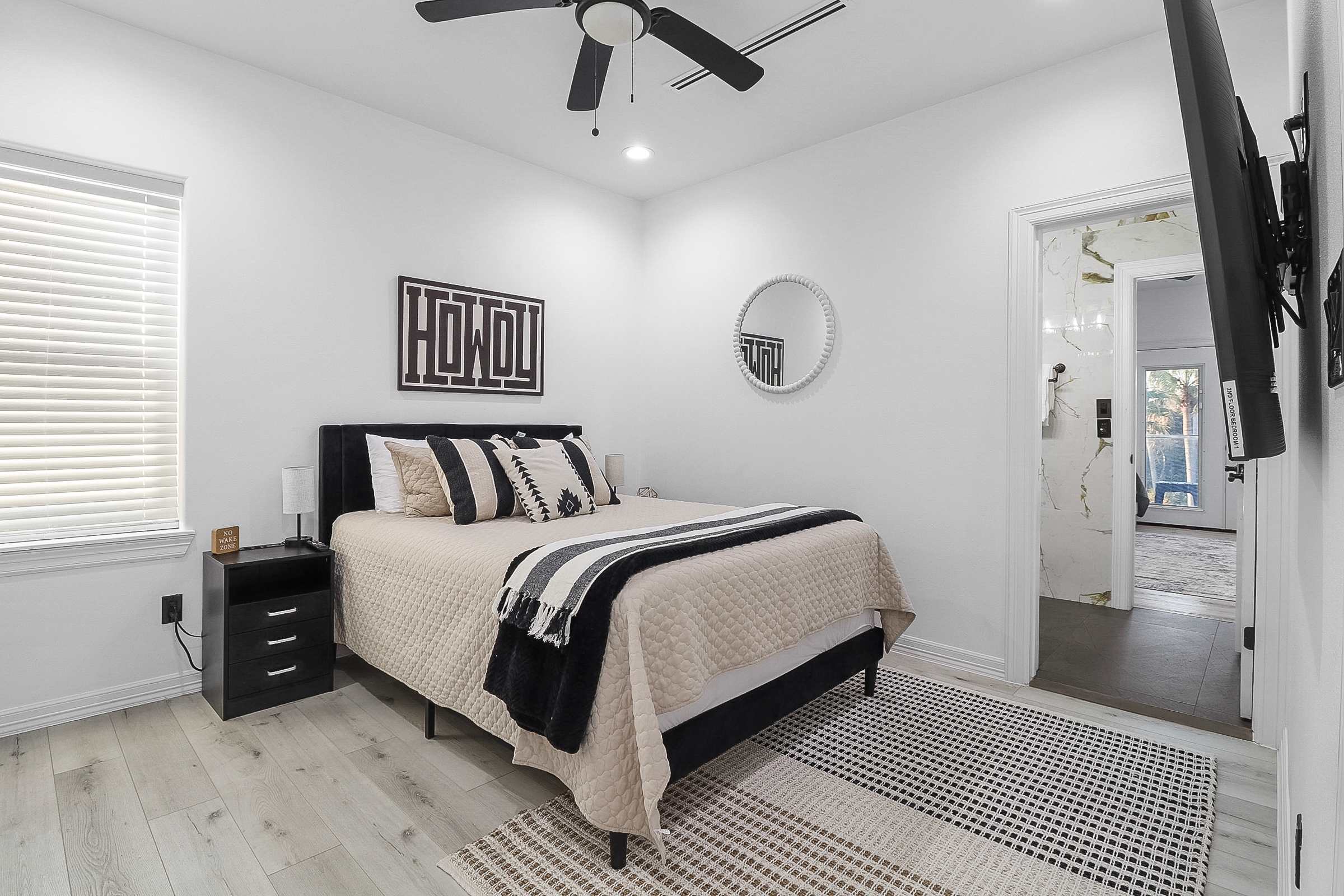 A modern bedroom with white walls, light wood-look flooring, a black upholstered bed with neutral bedding, a black nightstand, and a round white mirror, with a glimpse of a marble-tiled bathroom through an open doorway.