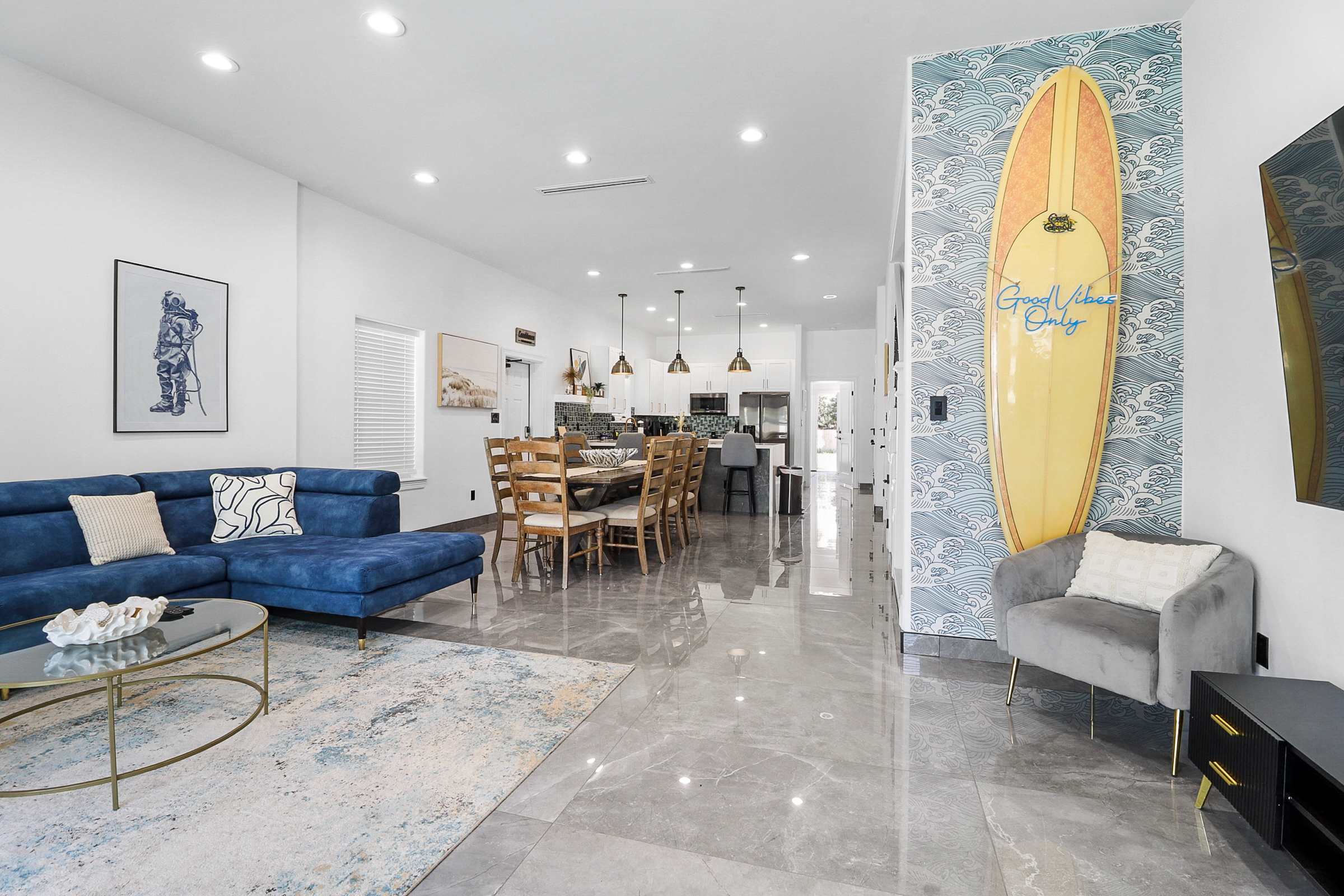 Open-concept living space with a blue sectional sofa, a glass coffee table, and a large surfboard against a blue wave-patterned accent wall. The view extends to a dining area and modern kitchen with white cabinetry.