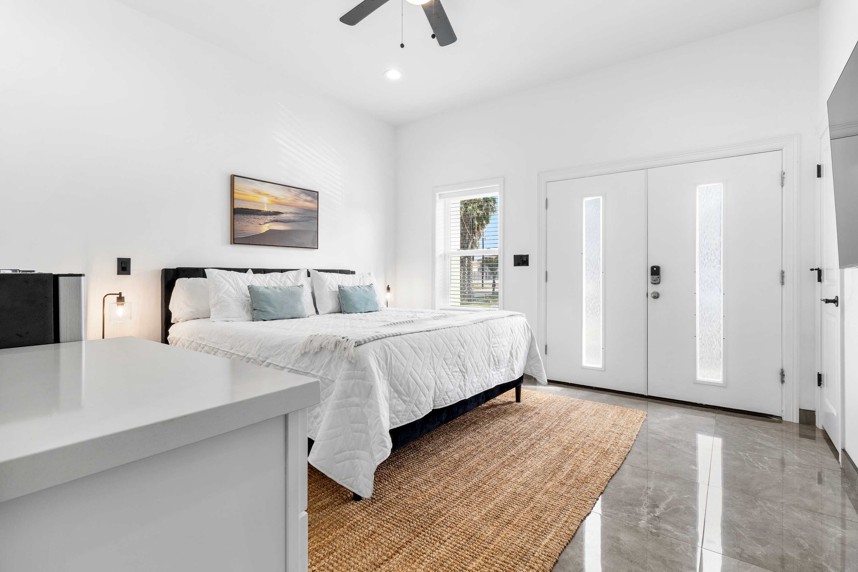 Wide shot of a bright bedroom with a king-size bed, a large jute rug, and double white entry doors with frosted glass panels, showcasing the modern white and grey aesthetic.