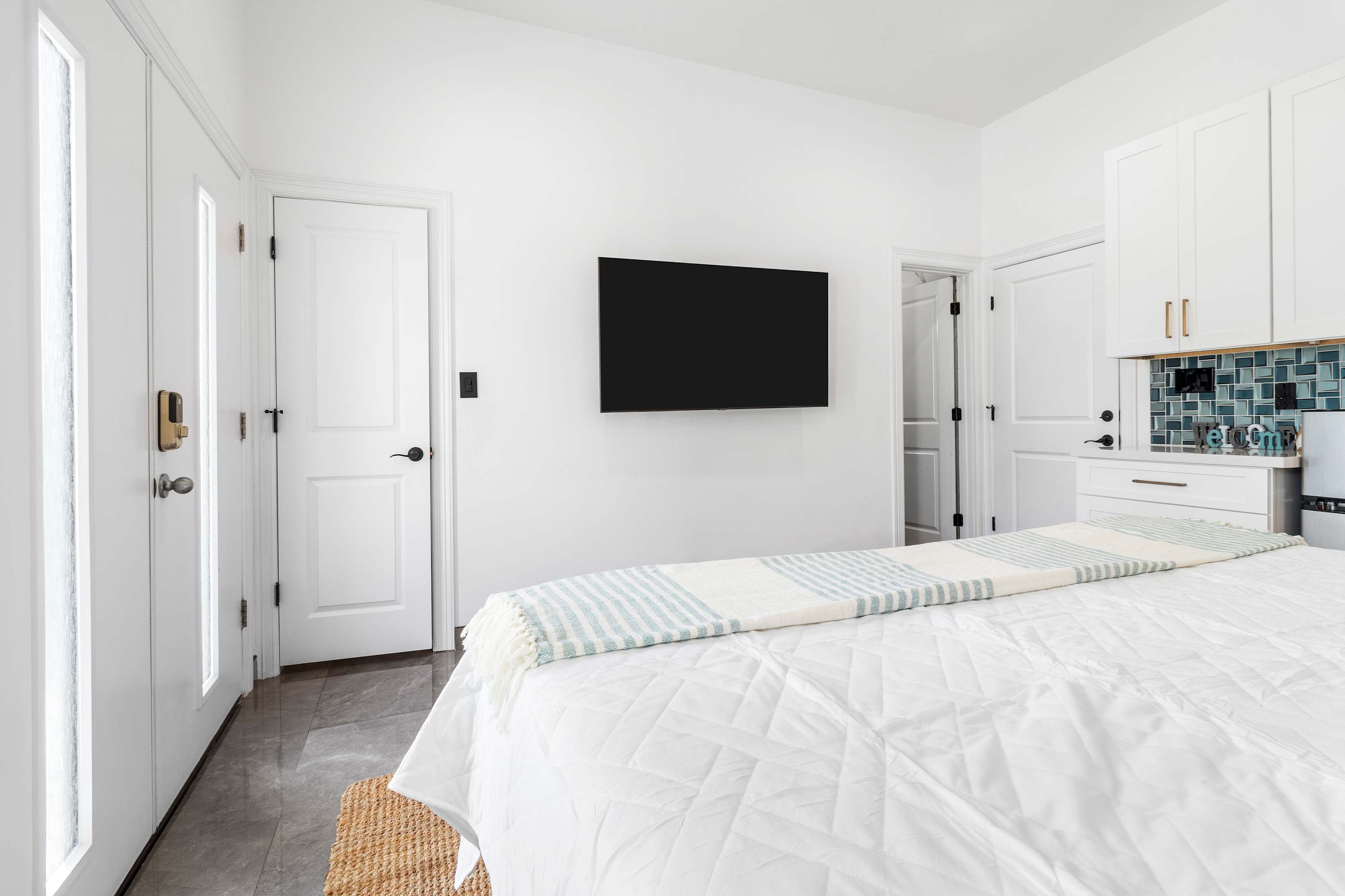 View of the bedroom from the foot of the bed, highlighting a wall-mounted flat-screen TV, white doors, and the continuation of the white quilted bedding with a striped throw.