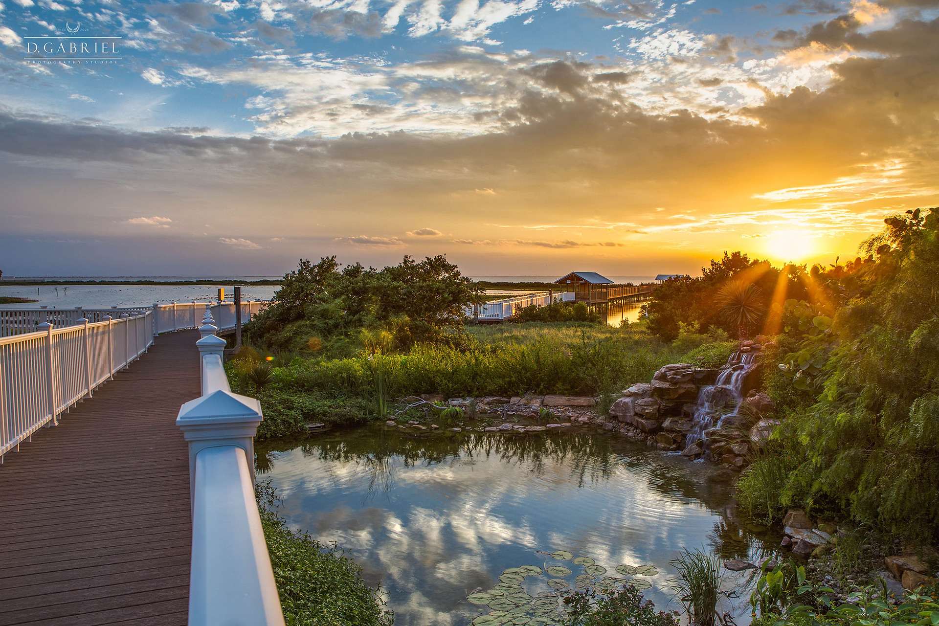 A picturesque sunset scene over a tranquil bay, featuring a wooden boardwalk with white railings extending into the distance, flanked by lush green foliage and a small rock waterfall. The sky is painted with warm orange and blue hues as the sun dips below the horizon.