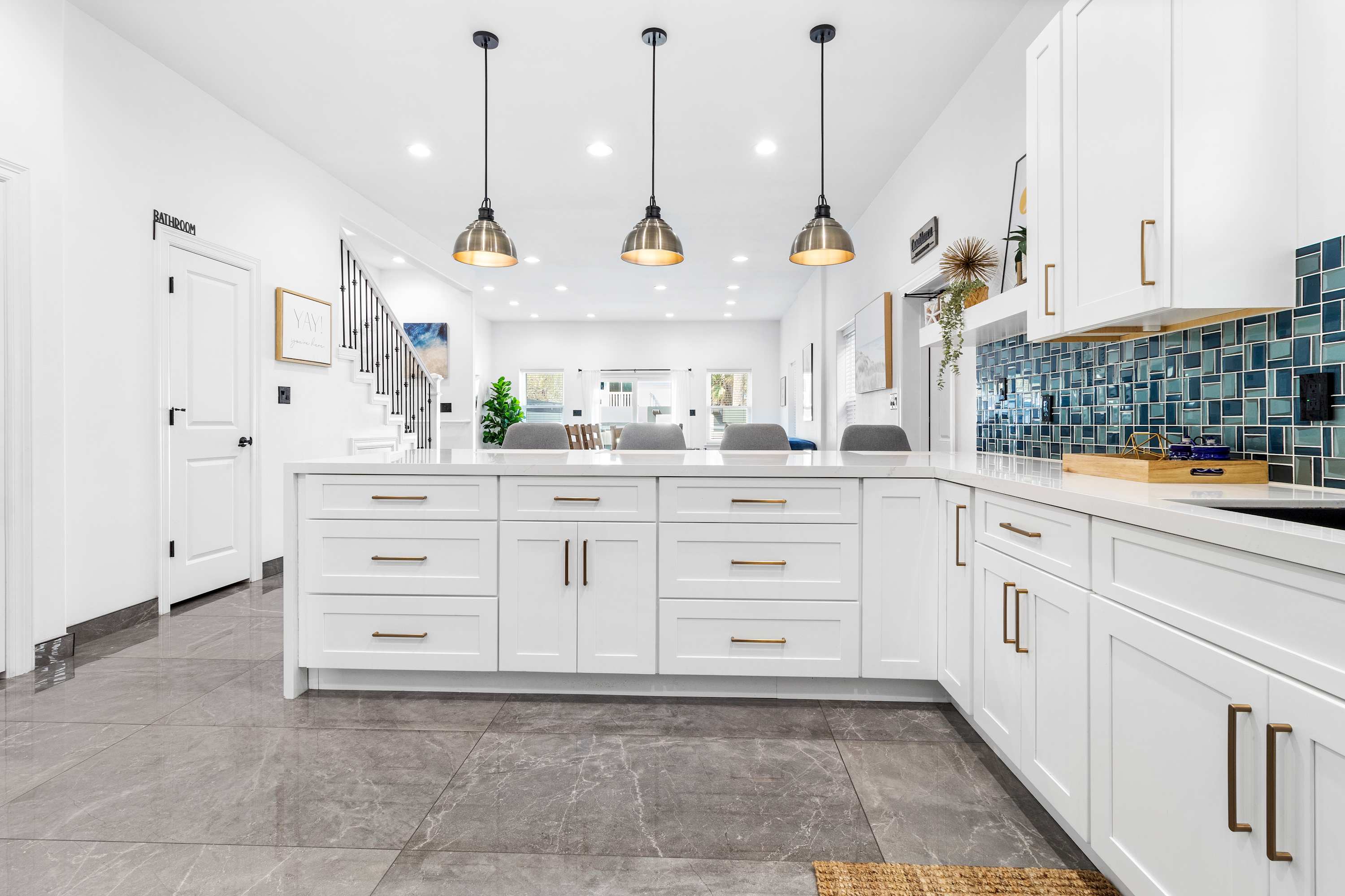 Contemporary kitchen island with white cabinetry and a white countertop, featuring three pendant lights overhead and seating for three. The open layout connects to the dining and living areas, showcasing the consistent grey tile flooring.
