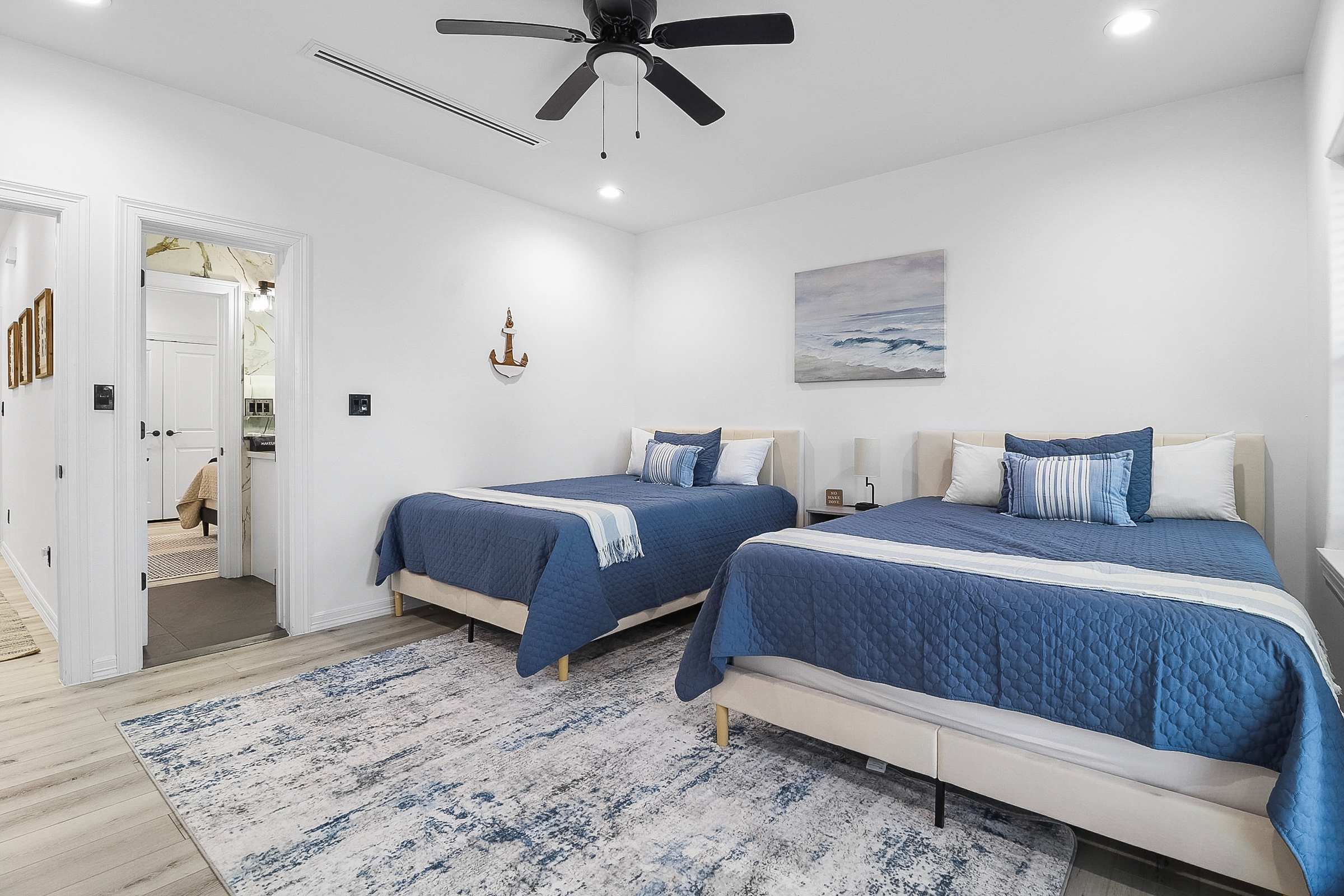 A bright bedroom with two queen beds featuring blue quilted bedding and decorative pillows, light wood-look flooring, white walls, and a blue and white abstract rug, with a glimpse of a bathroom through an open doorway.