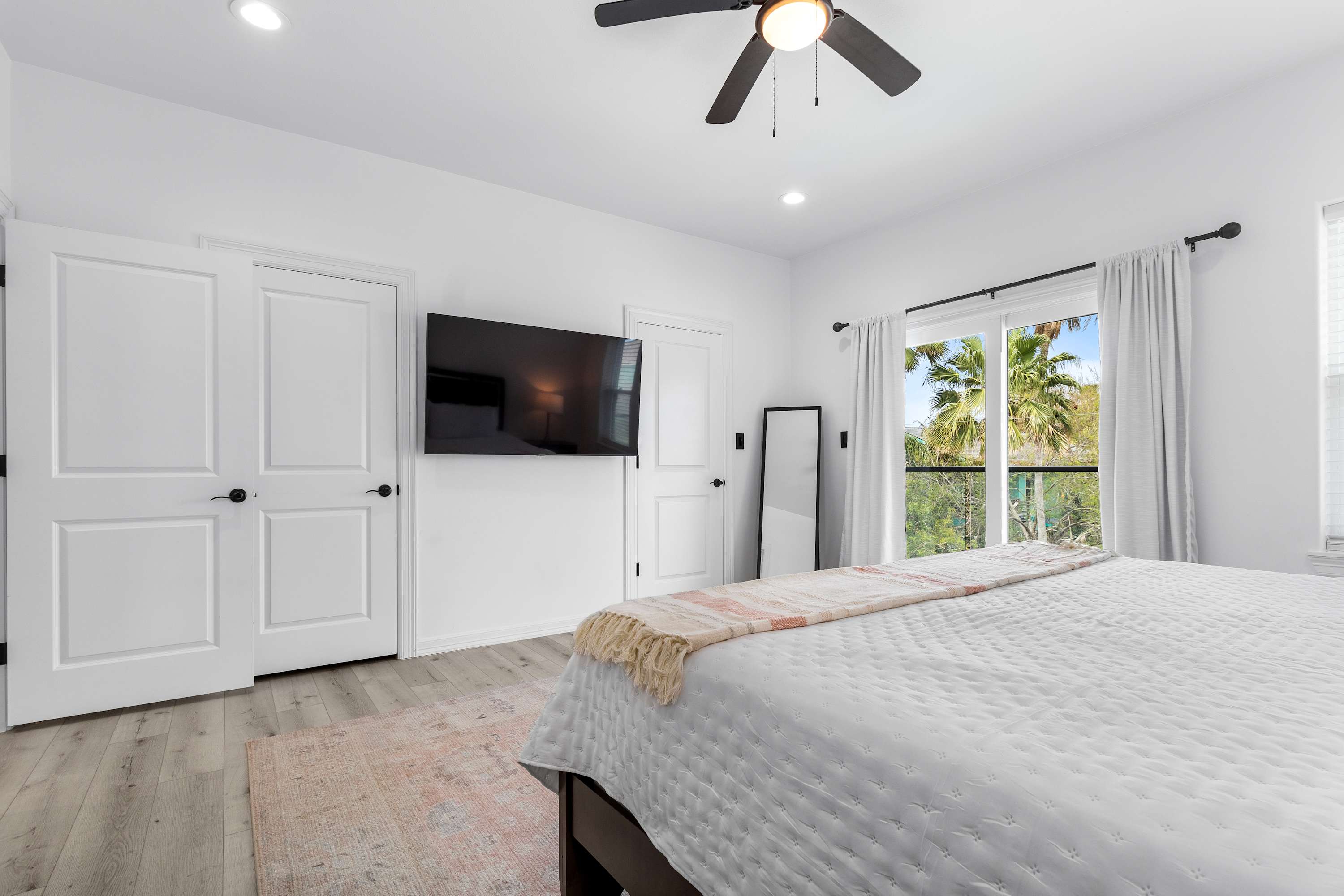 View of the bedroom showing a wall-mounted flat-screen TV, white double doors, and a large window with white curtains overlooking palm trees, providing a bright and airy feel.