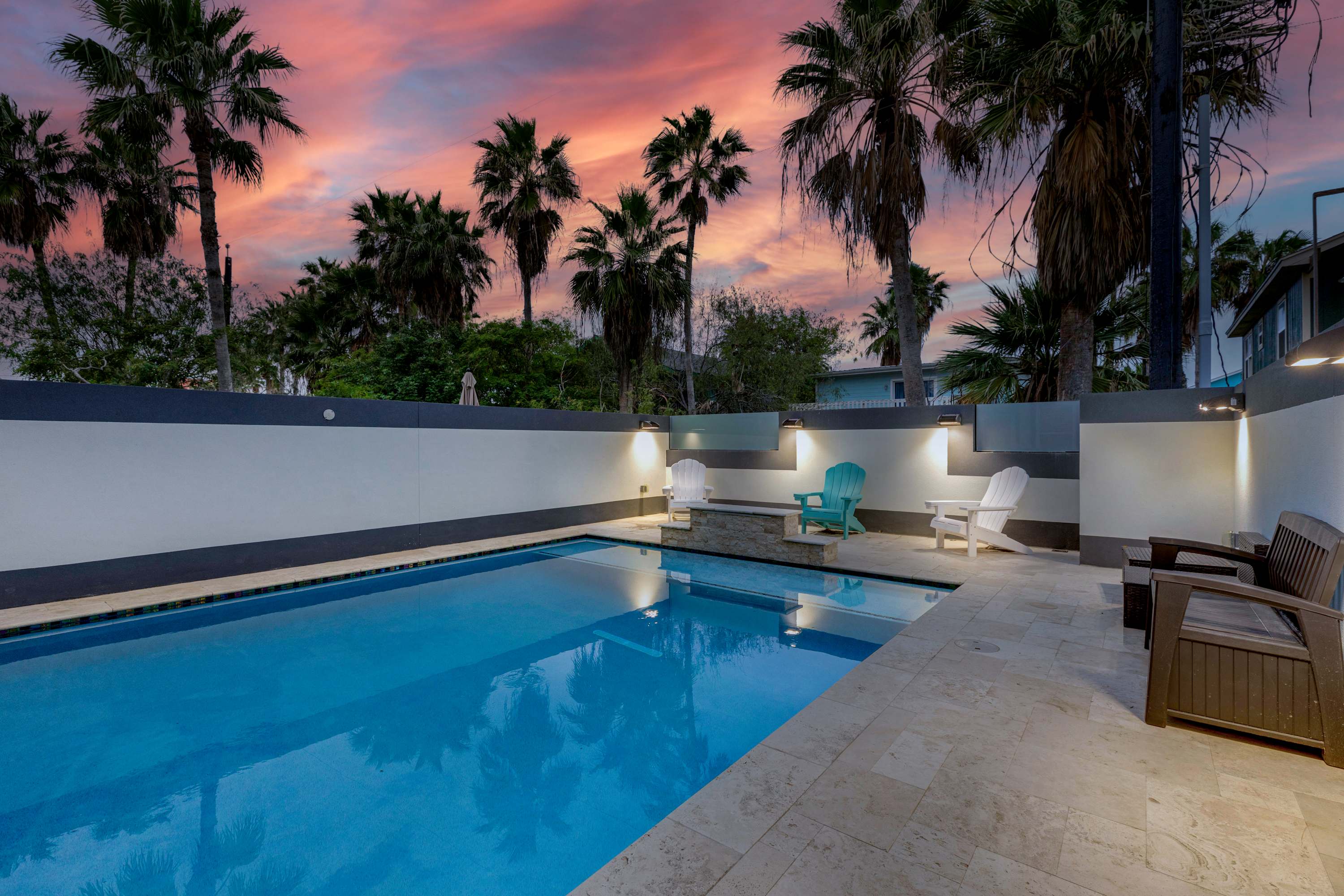 An inviting outdoor private pool area surrounded by light-colored paving, with several Adirondack chairs and lounge seating, illuminated by wall-mounted lights under a dramatic sunset sky with palm trees.