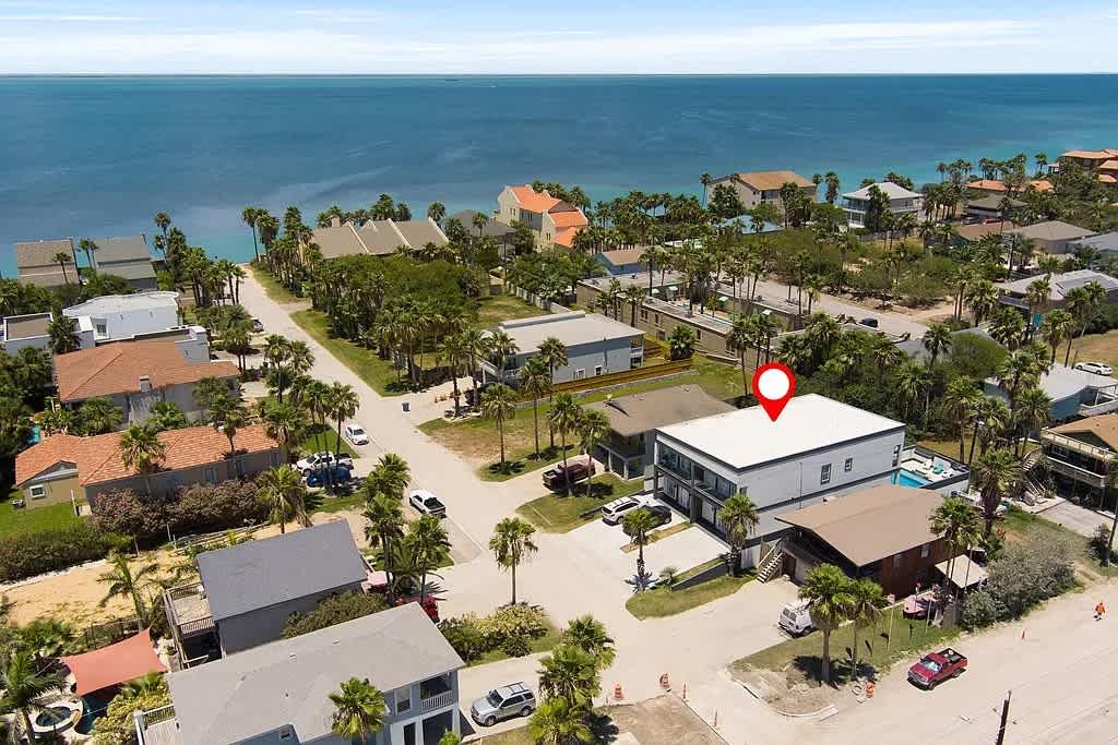 An aerial view of a coastal neighborhood showing the property (highlighted with a red pin) and surrounding houses, palm trees, and the expansive blue ocean or bay in the background under a bright sky.