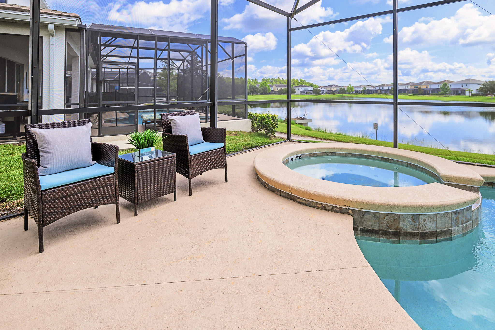 Screened-in outdoor area featuring a circular hot tub and two wicker chairs with blue cushions, overlooking a serene lake with houses in the distance, surrounded by green grass and a concrete patio.