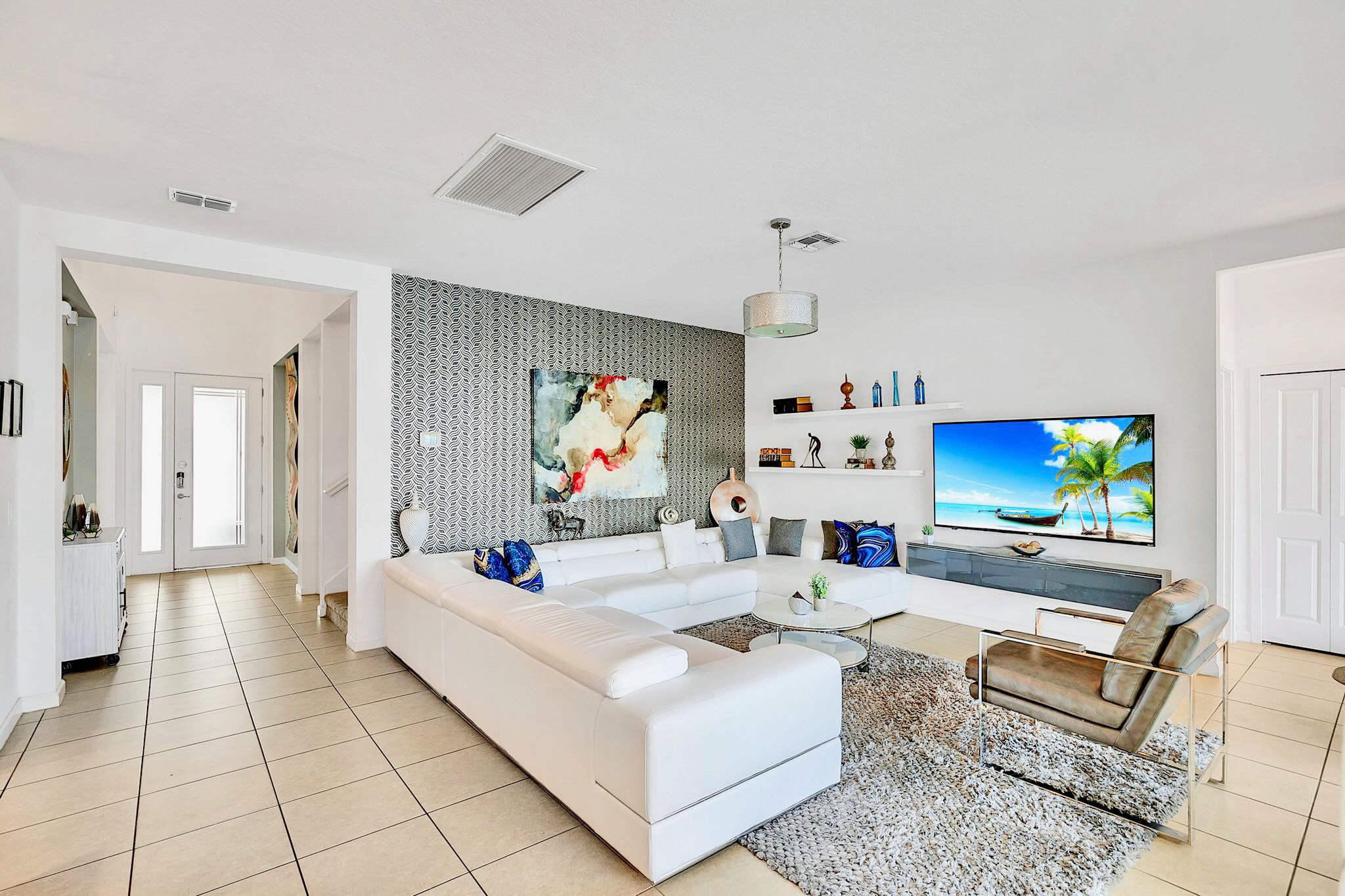 Spacious living room featuring a large white sectional sofa, a textured gray accent wall with abstract art, and a wall-mounted TV, all on light tile flooring.