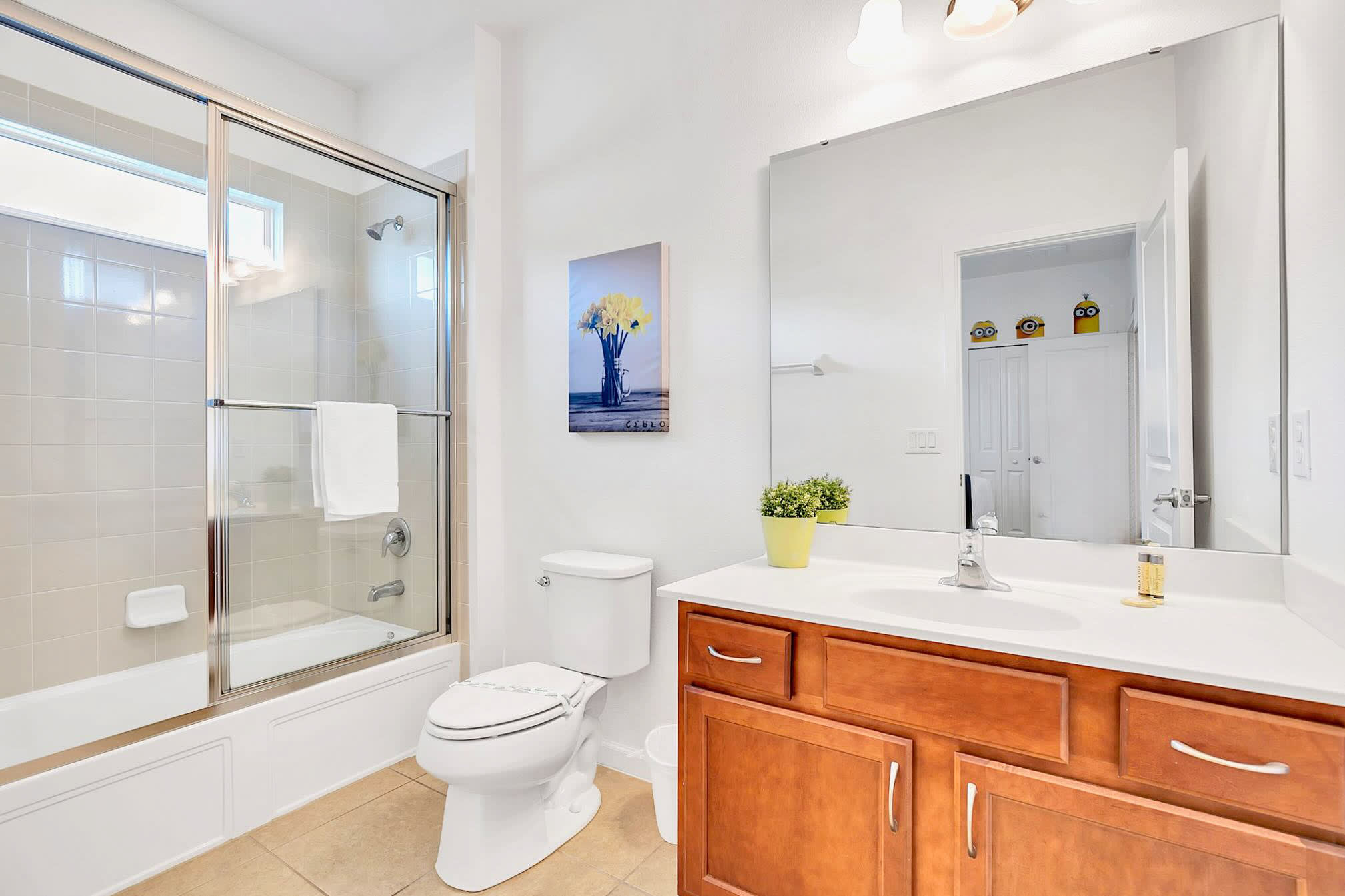 Bright bathroom featuring a white toilet, a wooden vanity with a white countertop and sink, and a shower-tub combo enclosed by a glass sliding door. A yellow floral art piece hangs above the toilet.