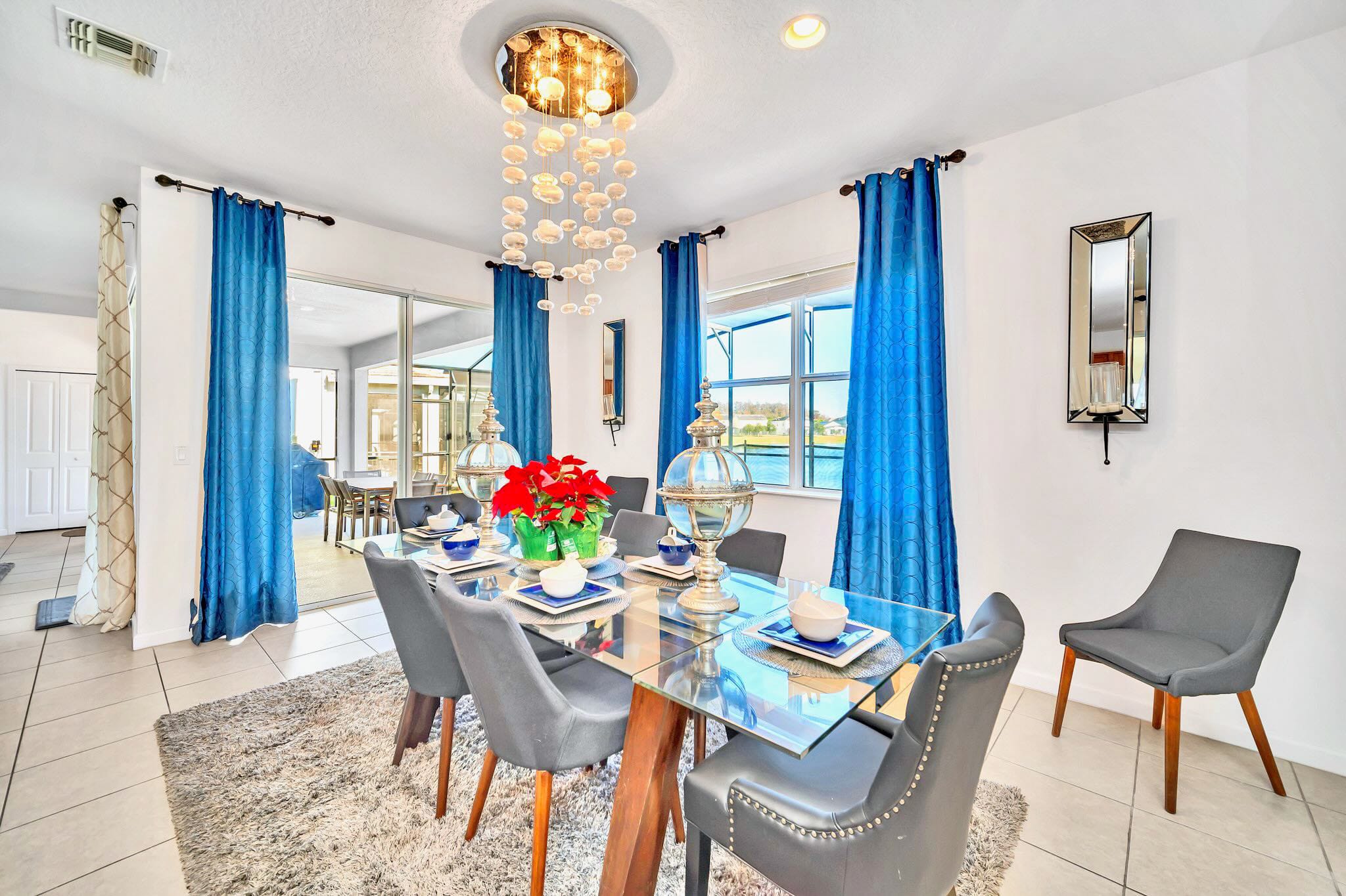 Elegant dining room featuring a glass-top table with gray upholstered chairs, a striking modern chandelier, and vibrant blue curtains framing windows with an outdoor view.