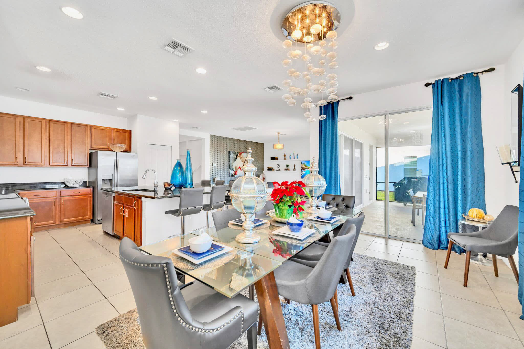 Elegant dining area with a glass-top table and gray upholstered chairs, illuminated by a unique bubble-like chandelier, adjacent to a modern kitchen with wooden cabinets and a breakfast bar, with sliding doors leading to an outdoor area.