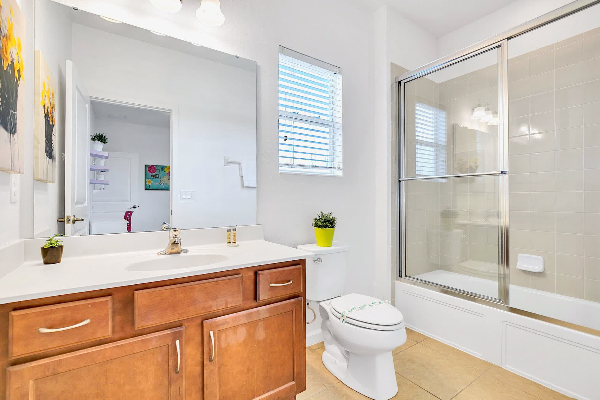 Modern bathroom with a wooden single vanity, white countertop, large mirror, toilet, and a shower/tub combo with a sliding glass door and light tiled walls.