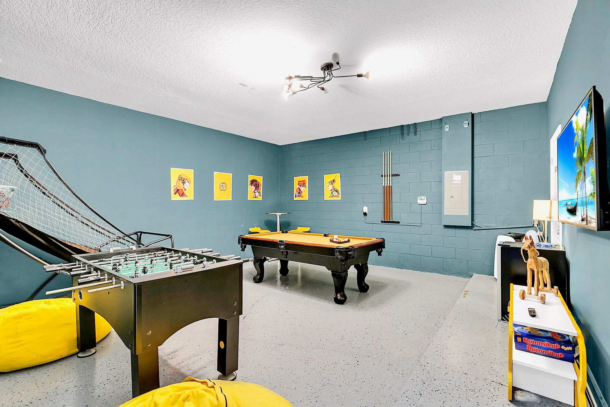 Vibrant game room featuring a pool table with a yellow felt, a foosball table, and a basketball arcade game, set against teal blue walls and a light gray speckled floor.