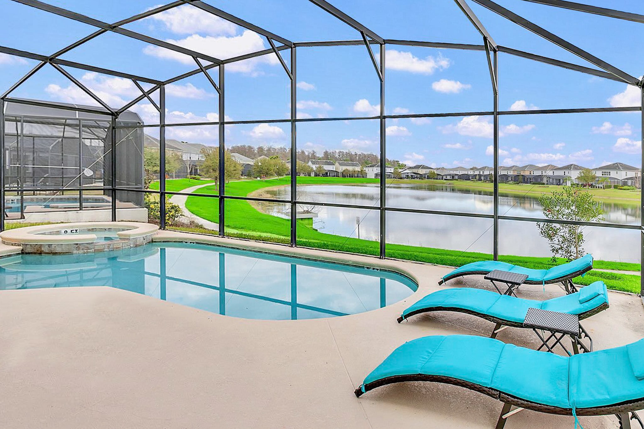 Screened-in outdoor pool area with a kidney-shaped pool and integrated hot tub, surrounded by light concrete decking and several blue lounge chairs, offering expansive views of a lake and distant houses under a blue sky.